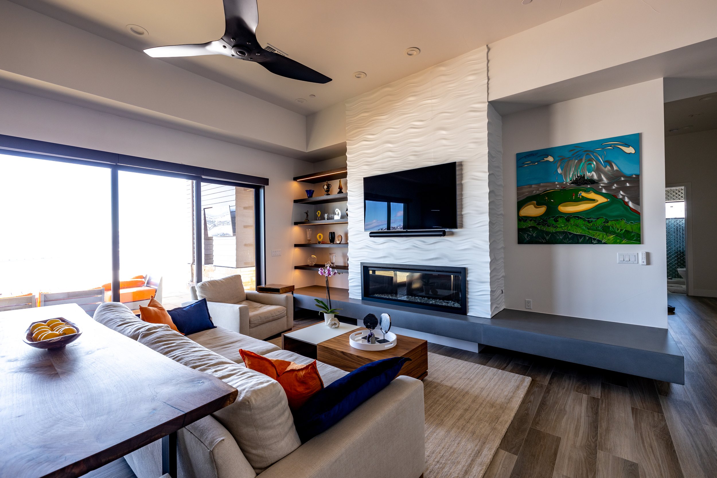 Modern living room with a wall-mounted TV above a fireplace, white textured accent wall, large window with sliding doors, beige sofa with orange and navy pillows, wooden coffee table with decorative objects, colorful abstract painting on the wall, bu