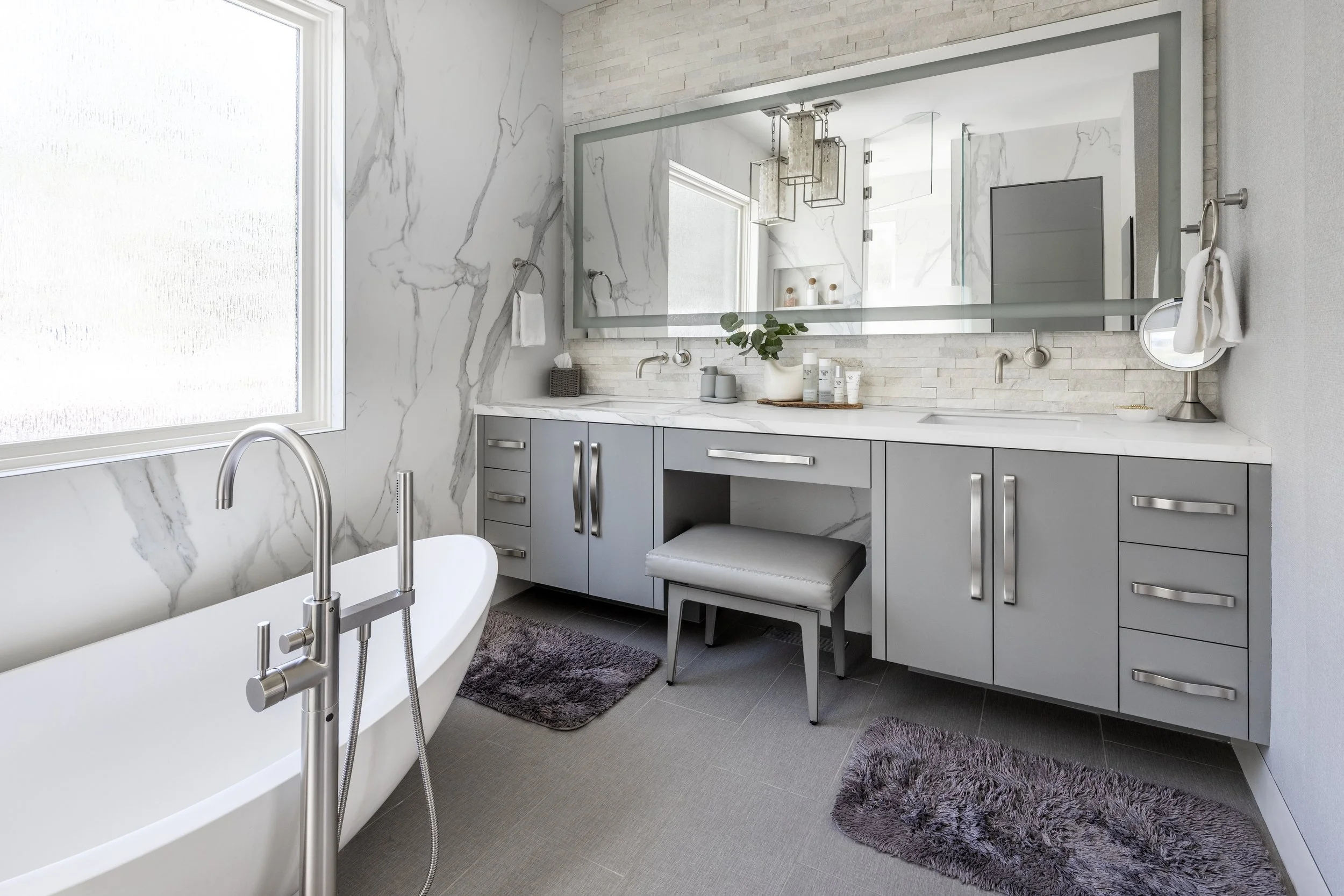 Mountain modern bathroom with luxe freestanding tub seated on top of hydronic heated porcelain grey linen tiles and crystal rock pendants suspended above. Walls are clad in porcelain slabs that look like a Calcutta marble. Custom painted vanity incor