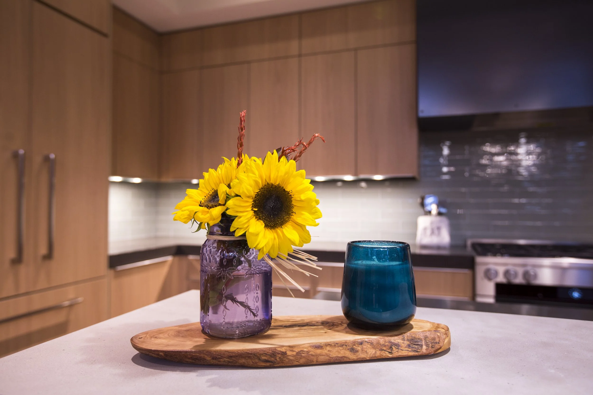 Yellow sunflowers in a purple glass jar and a blue glass tumbler on a wooden tray in a modern kitchen.