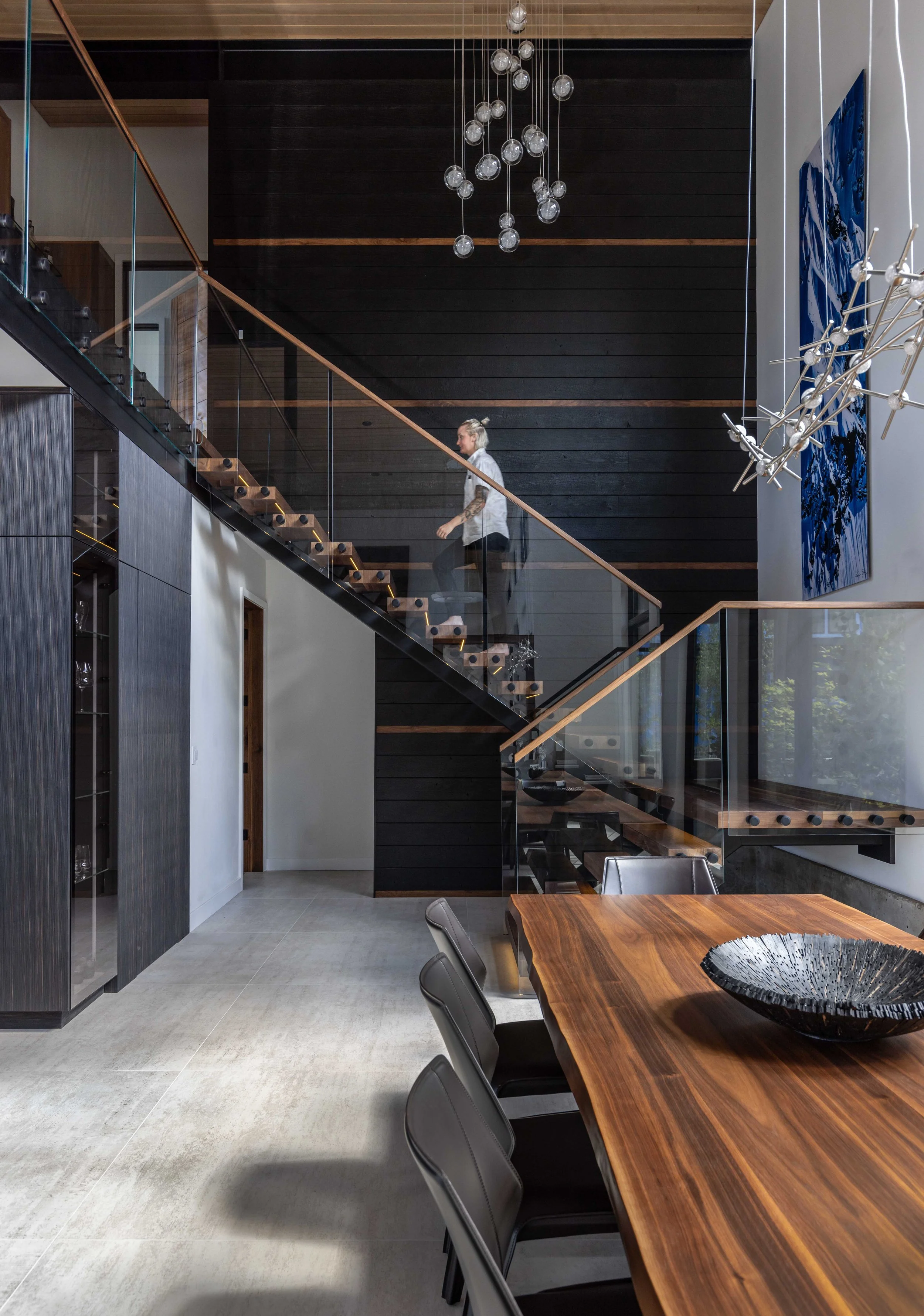 Modern dining area with a large wooden table, dark chairs, and a black decorative bowl. The staircase with glass railings and wooden steps leads up to a second floor. The space features a black accent wall, artwork, and contemporary light fixtures.