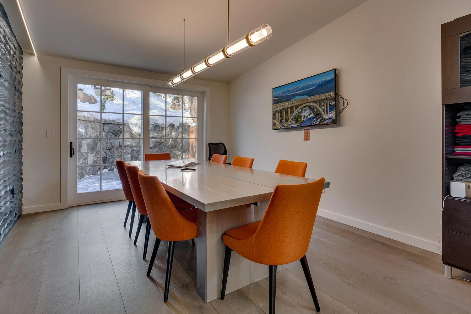 Indoor dining room with a large wooden table surrounded by eight orange chairs, sliding glass door showing snow-covered exterior, mountain scenery on television, and modern decor.
