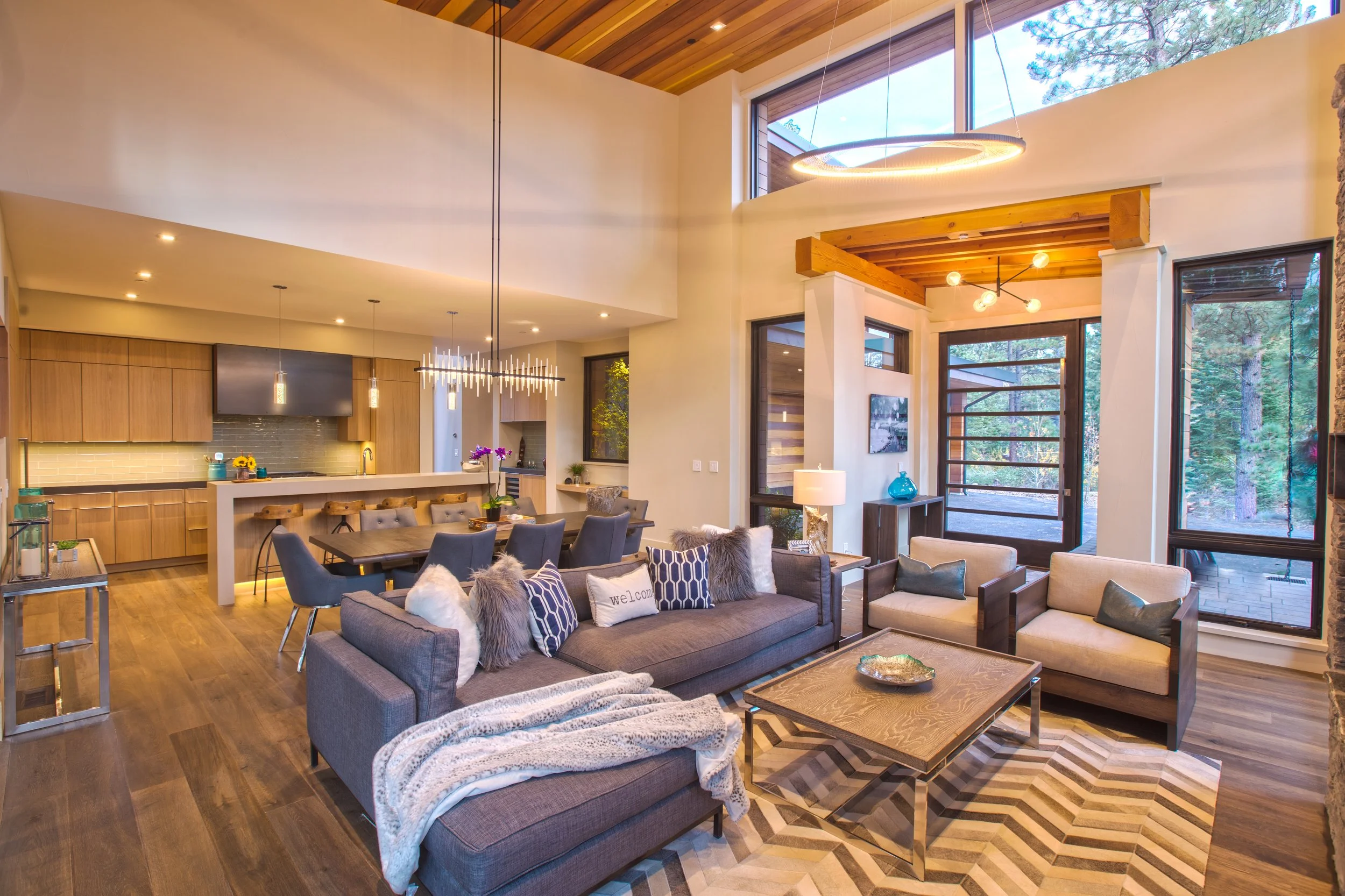 Open-concept living area and kitchen with wood floors, high ceilings, modern furniture, large windows, and wooden accents.