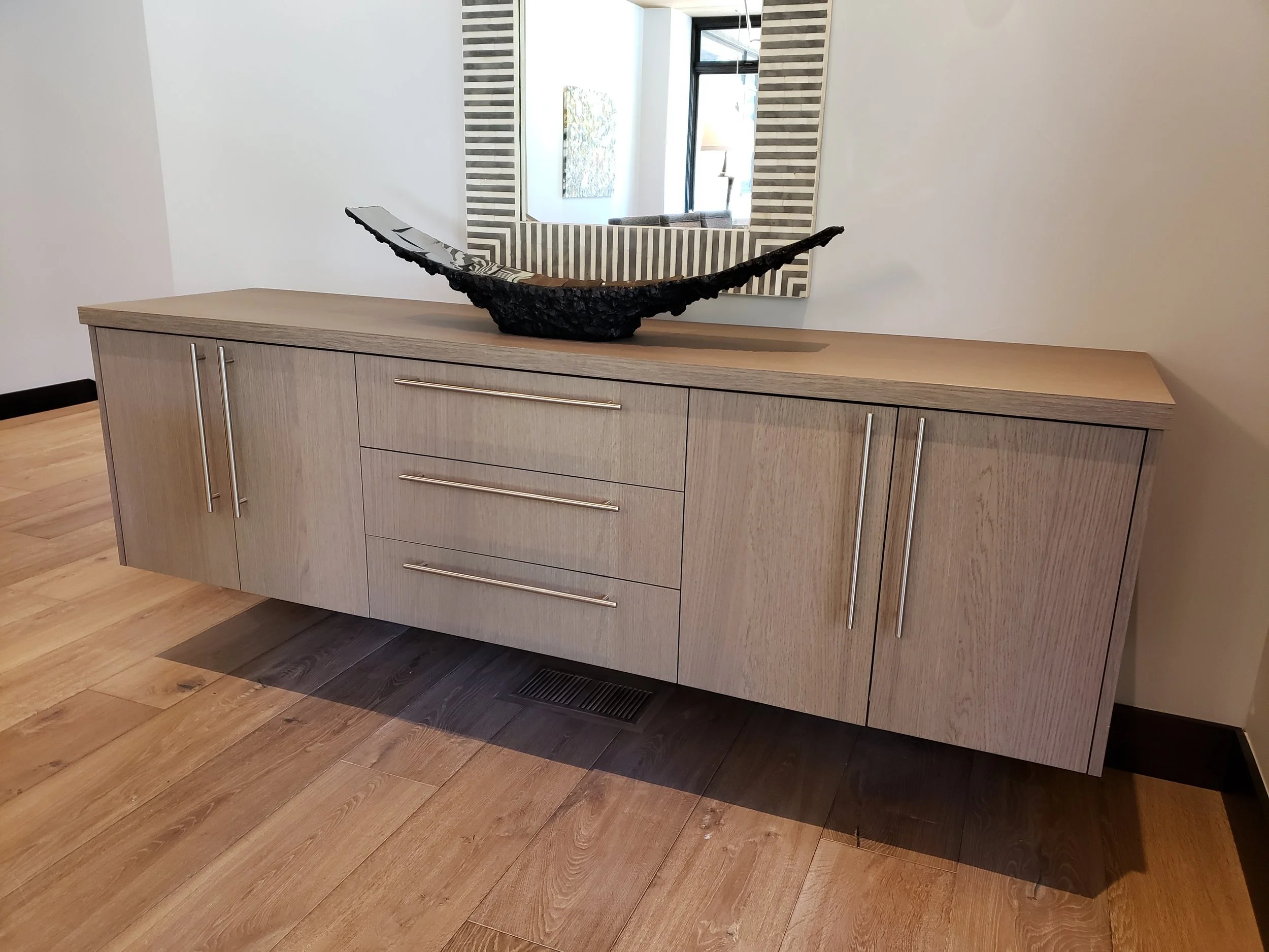 A custom floating wood sideboard with chrome handles, topped with a black decorative bowl, placed against a white wall with a large mirror reflecting a window and part of a living room. Designed by Emily Roose.