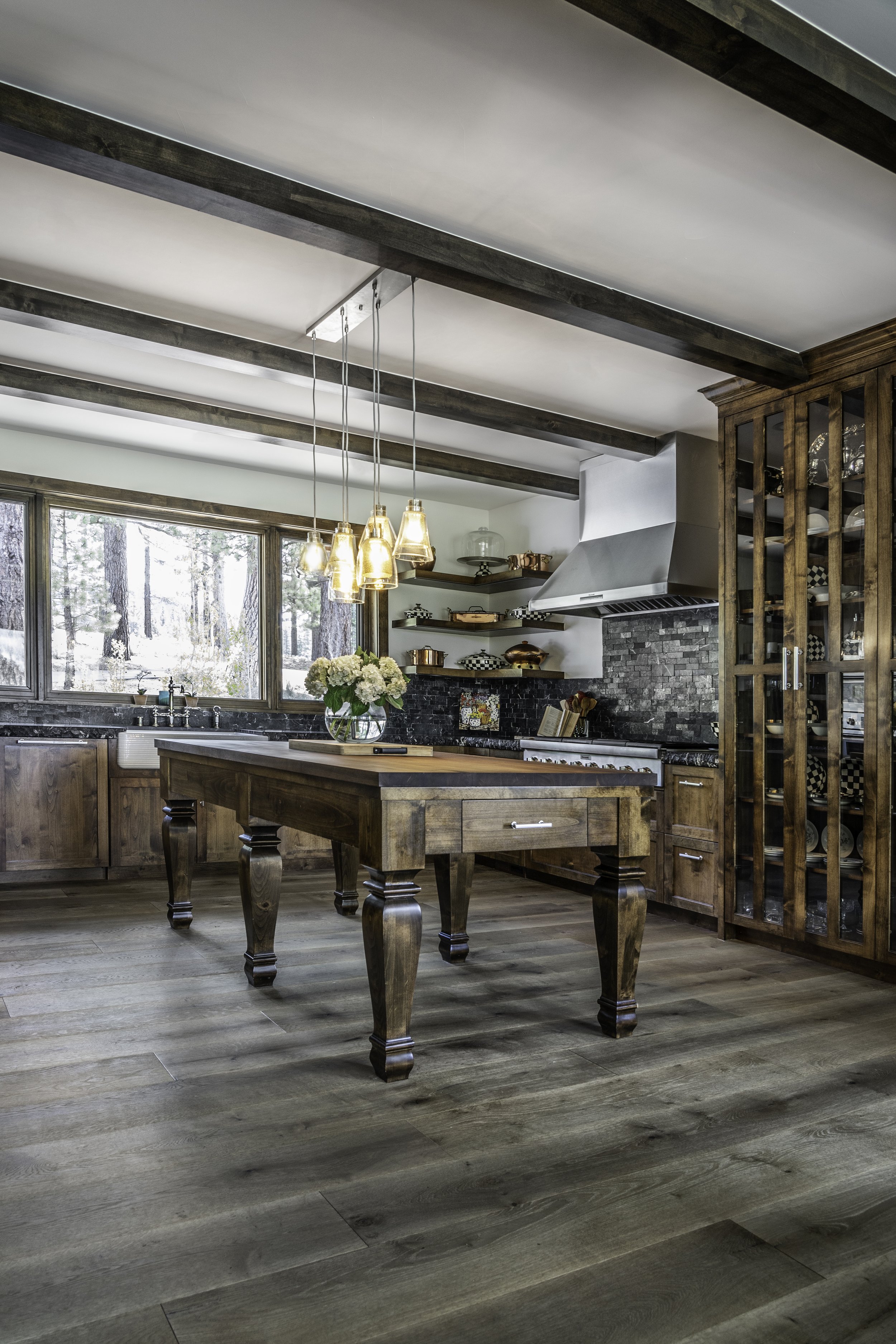 A rustic kitchen with wooden cabinets, a kitchen island with a vase of flowers, modern pendant lights, large windows showing trees outside, a stove with a vent hood, and shelves with copper and patterned dishes.