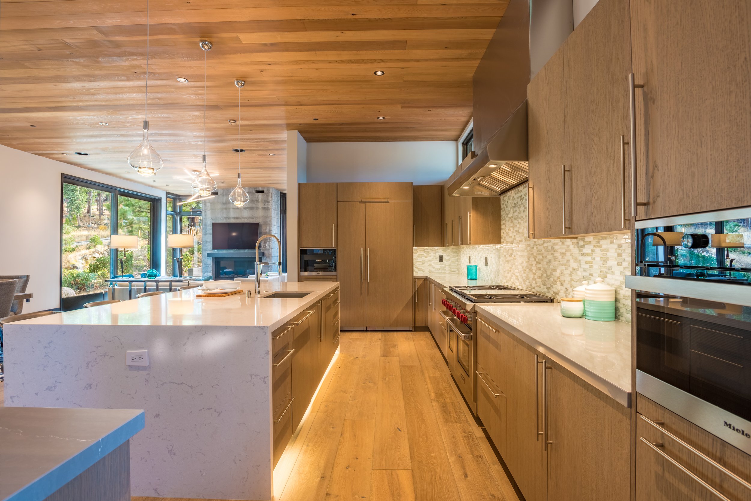Modern kitchen with wood cabinets, marble island, and pendant lights, overlooking a living room with a stone fireplace and large windows showing greenery.