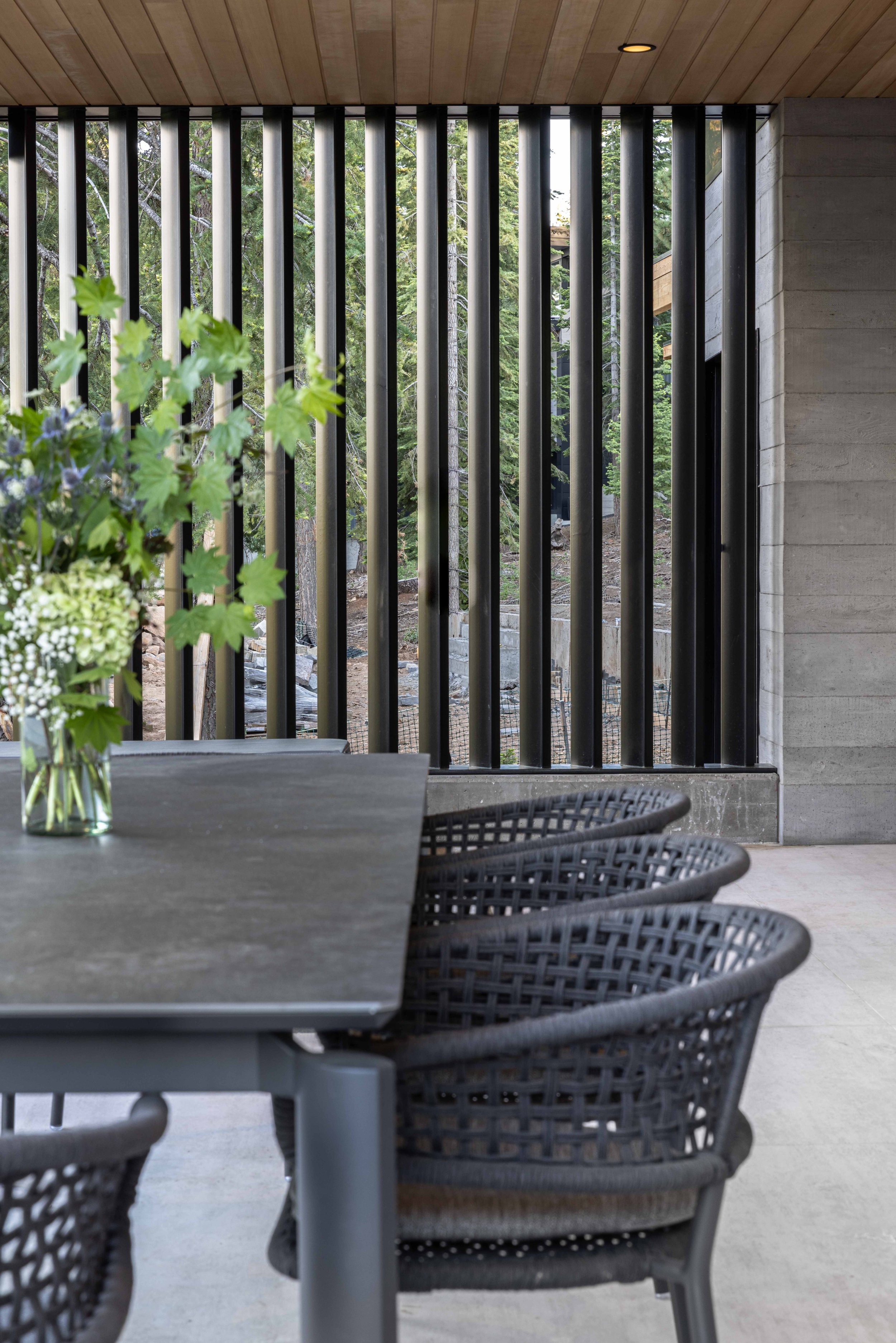 Interior of a modern dining area with a table, woven black chairs, a vase with flowers, and vertical black metal bars outside revealing trees.