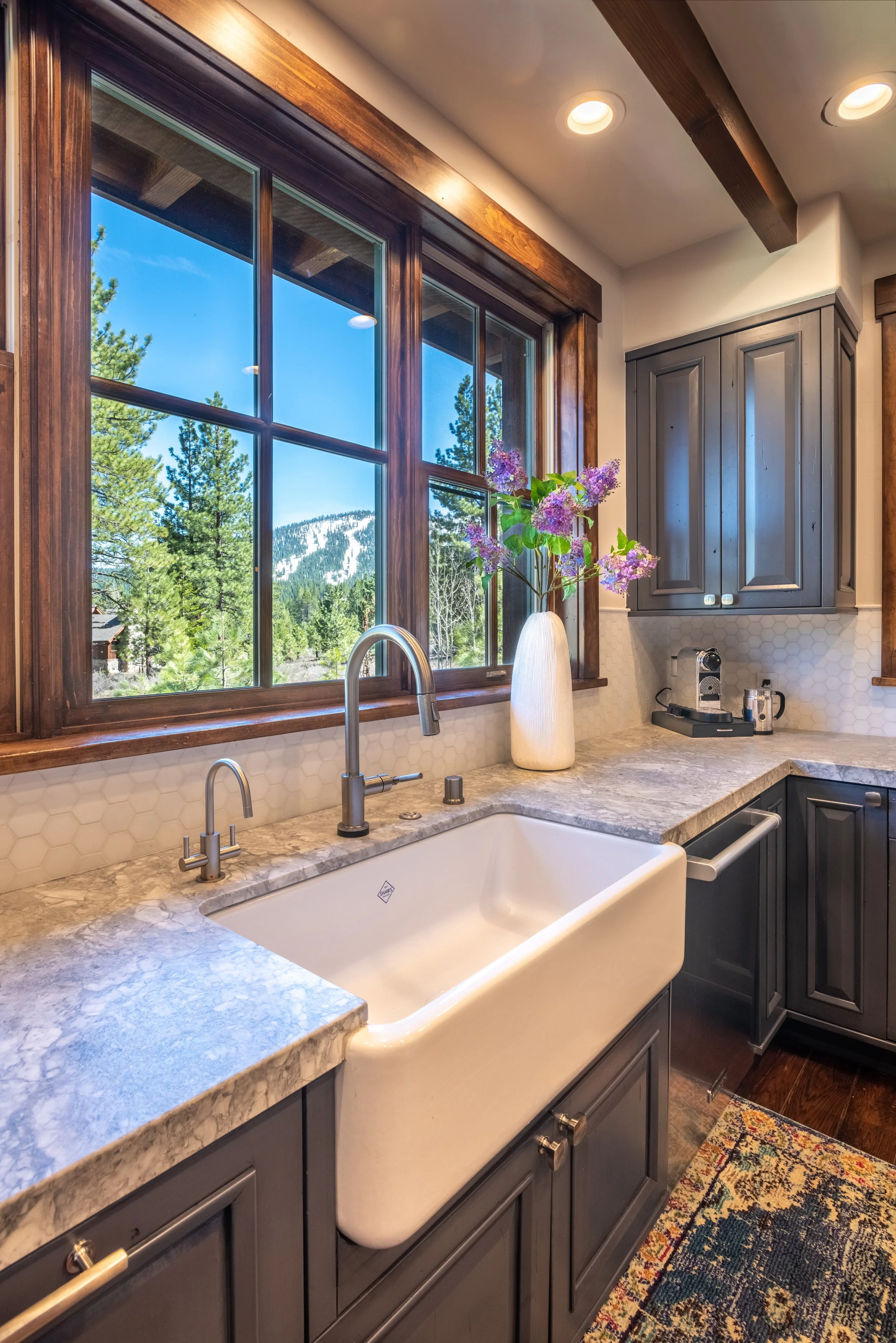 A kitchen with a farmhouse sink, granite countertops, grey cabinets, a vase with purple flowers, a window with a mountain view, and a colorful rug.