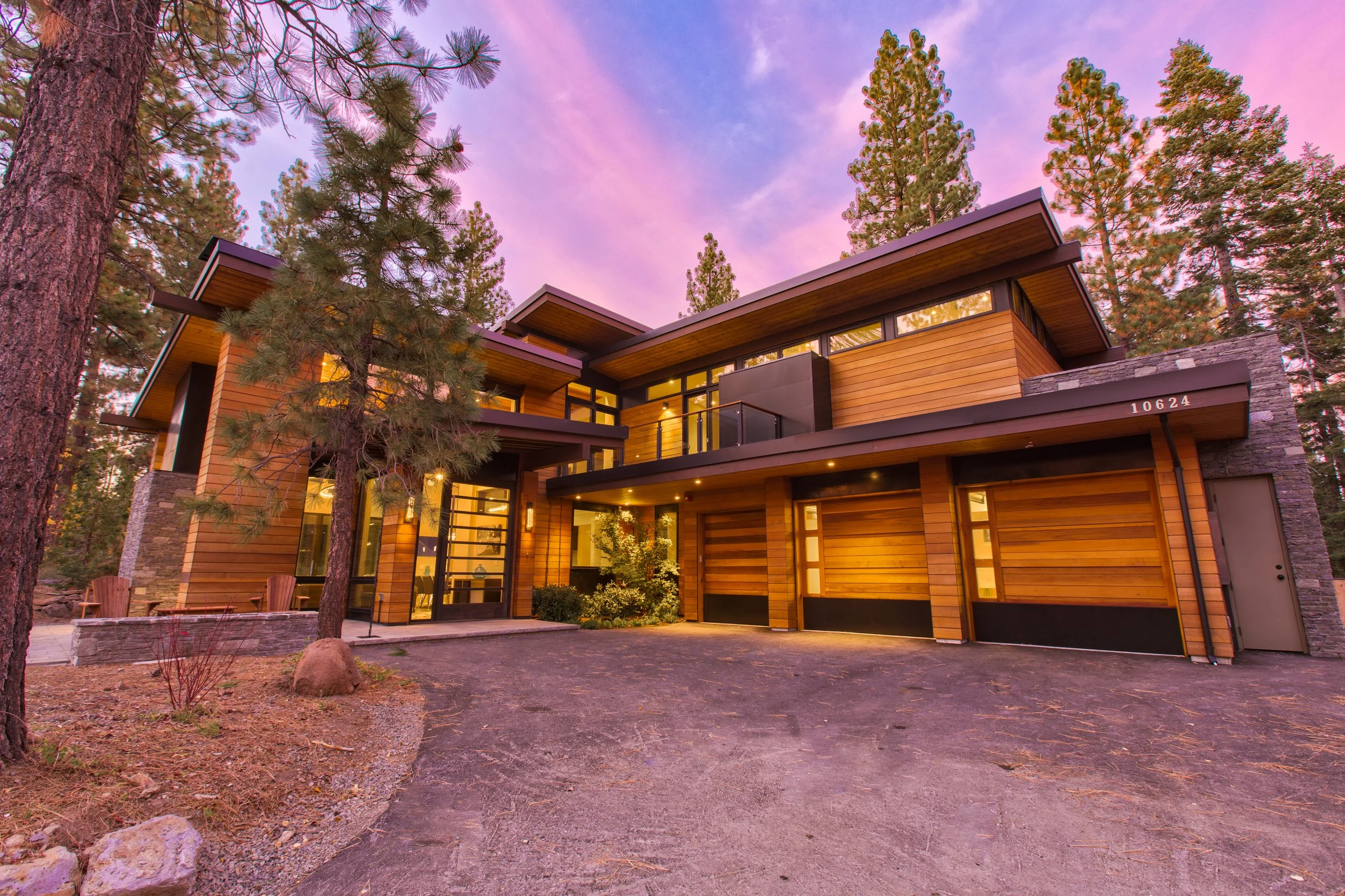 Modern wooden house with a large driveway surrounded by tall pine trees, under a pink and purple sky at sunset.