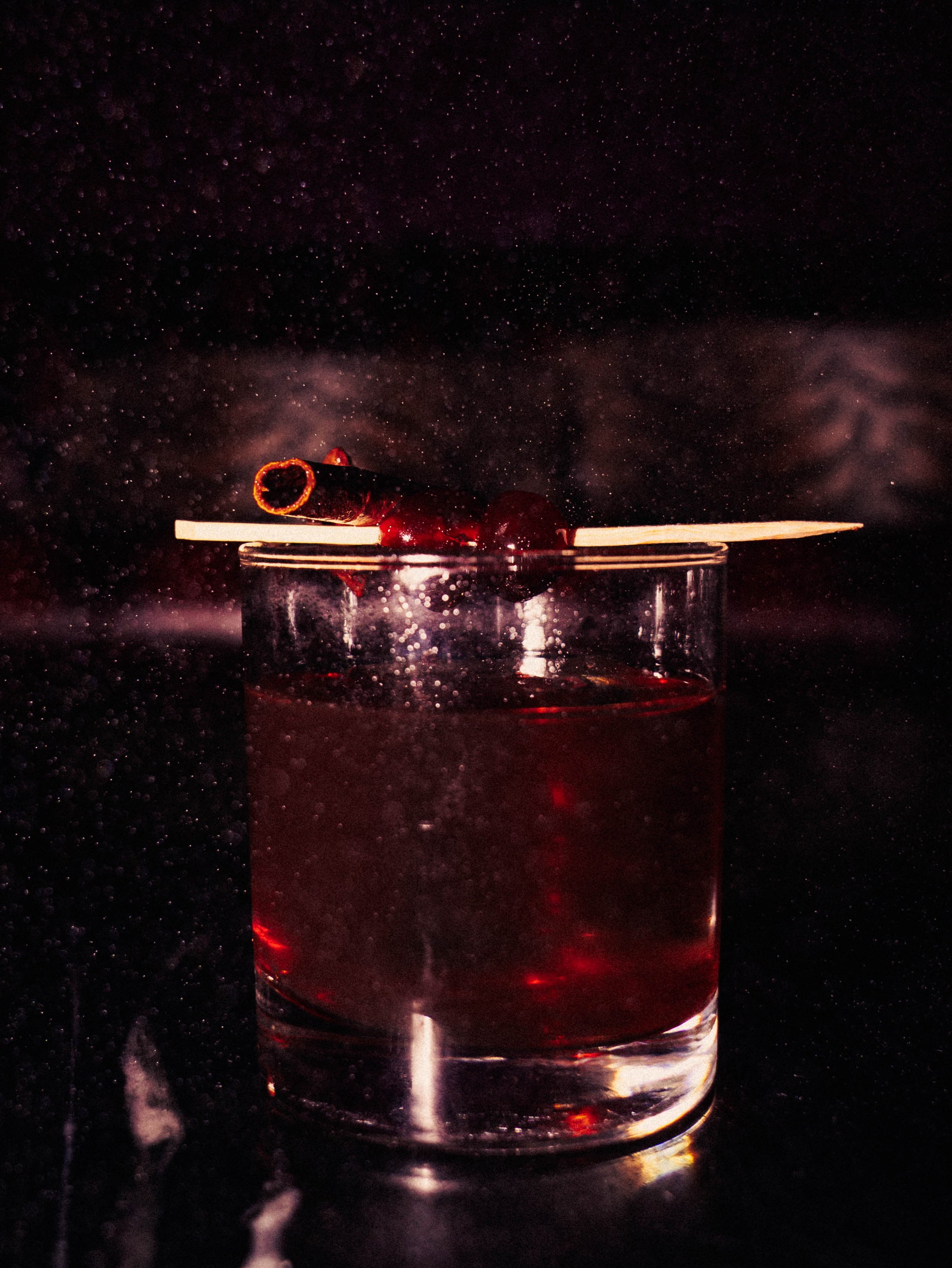 A clear glass filled with a dark red drink, garnished with a cherry and a cinnamon stick, against a dark background with droplets or particles.