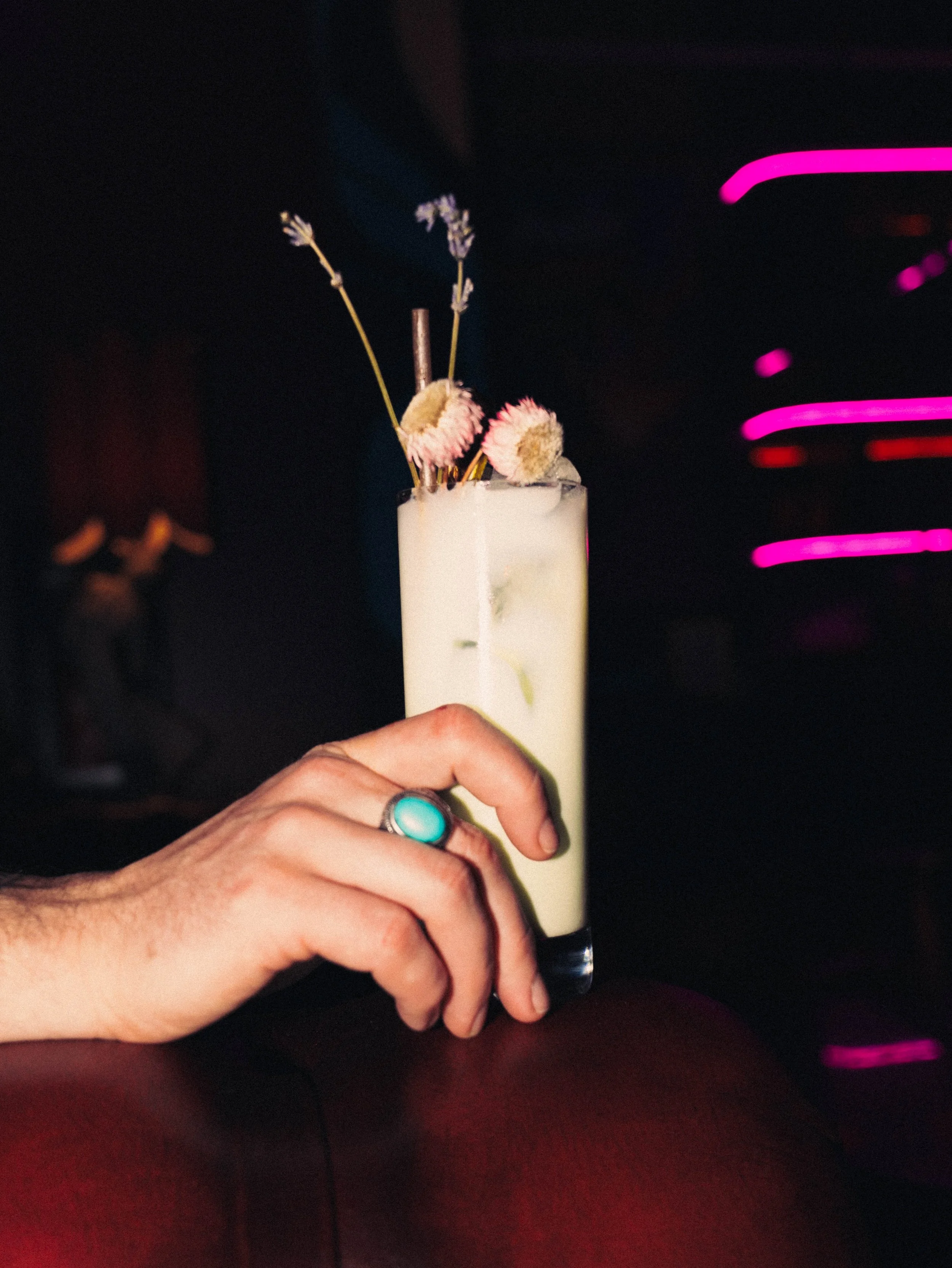 A person's hand holding a tall glass of cocktail with flower garnishes against a dark background with pink neon lights.