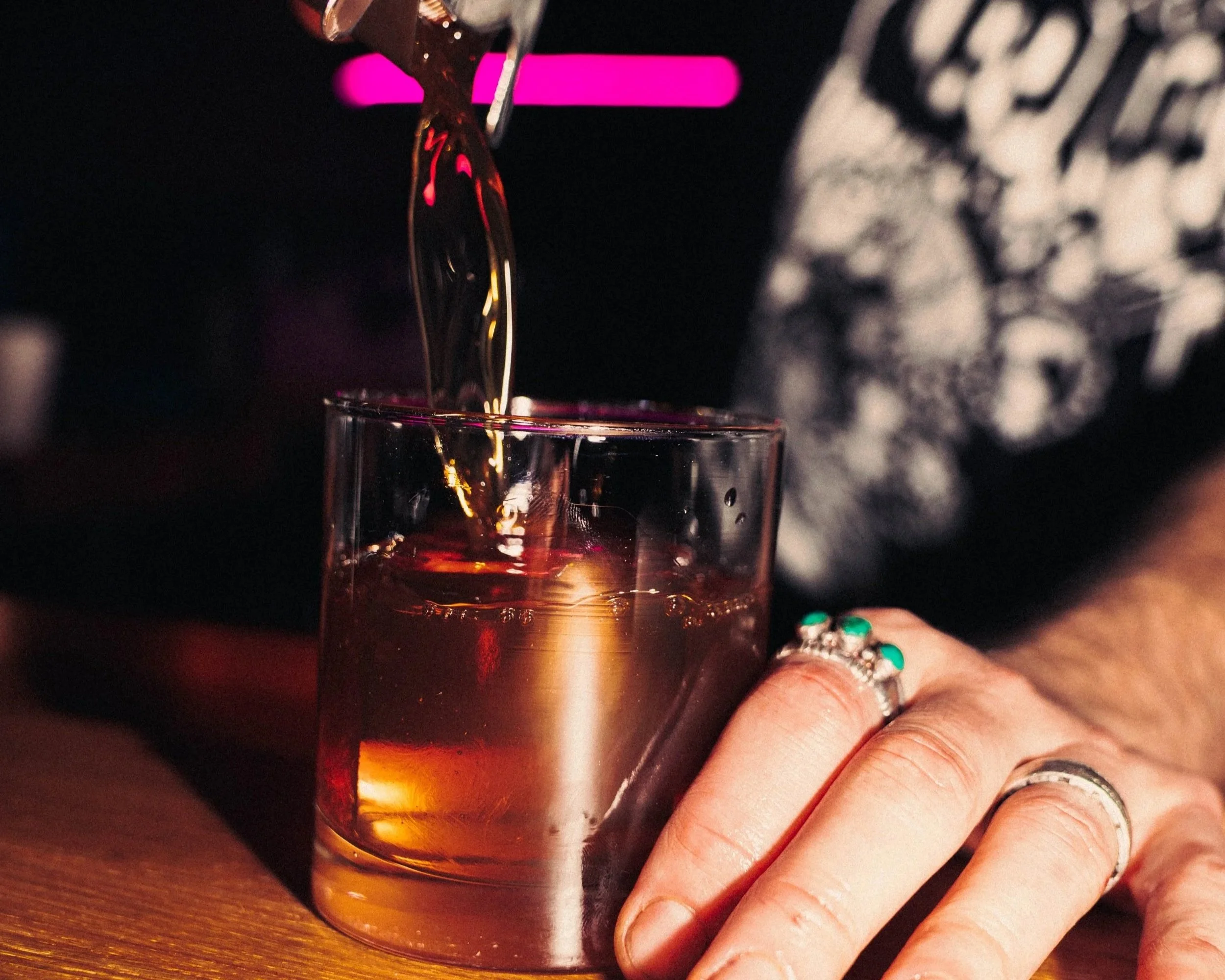 A person pours a golden-colored beverage from a bottle into a clear glass on a wooden surface, with a person wearing rings and a black patterned shirt nearby.