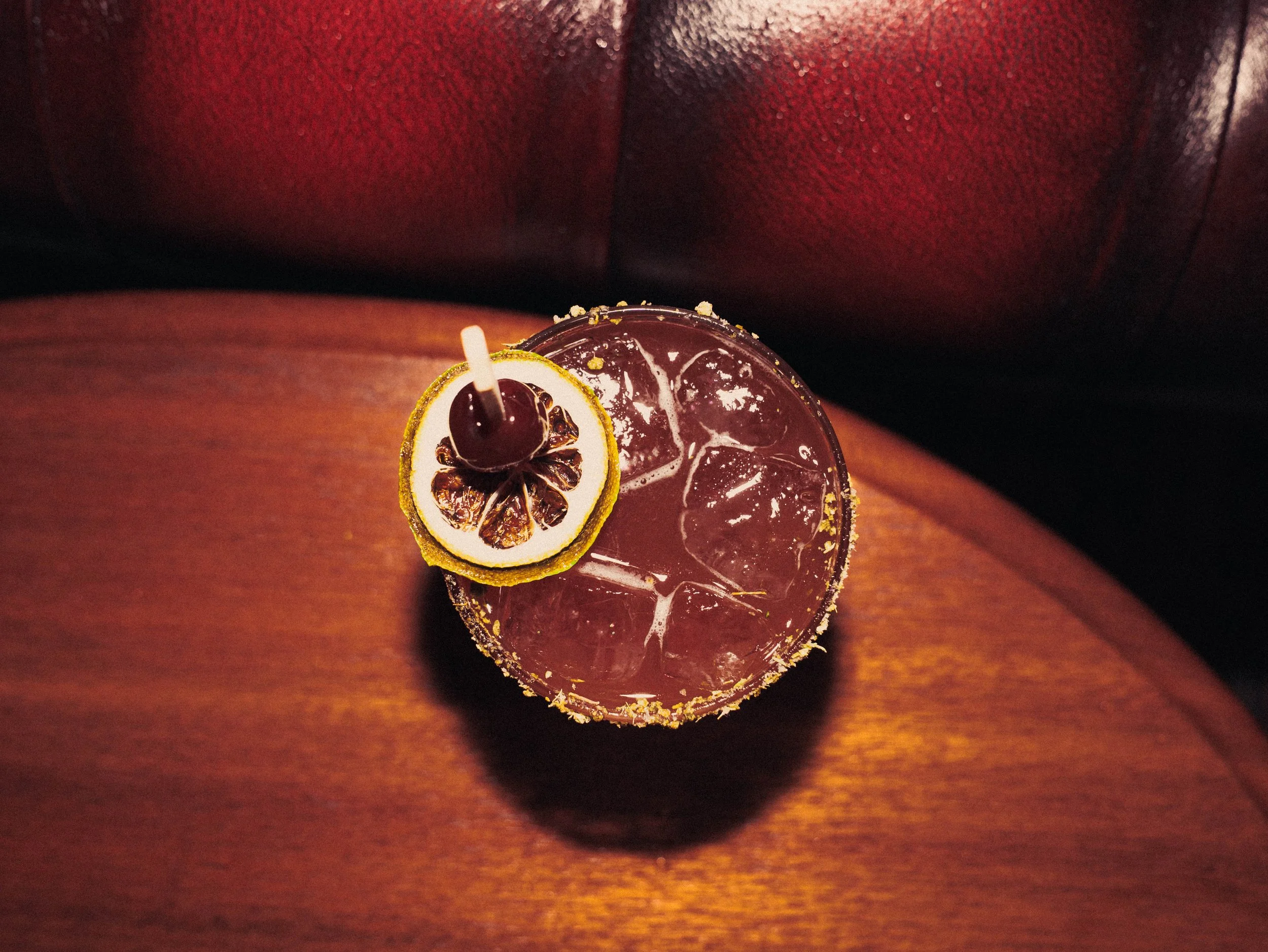 Top-down view of a cocktail on a wooden table with ice cubes, a slice of lemon, and a cherry garnished on the rim.
