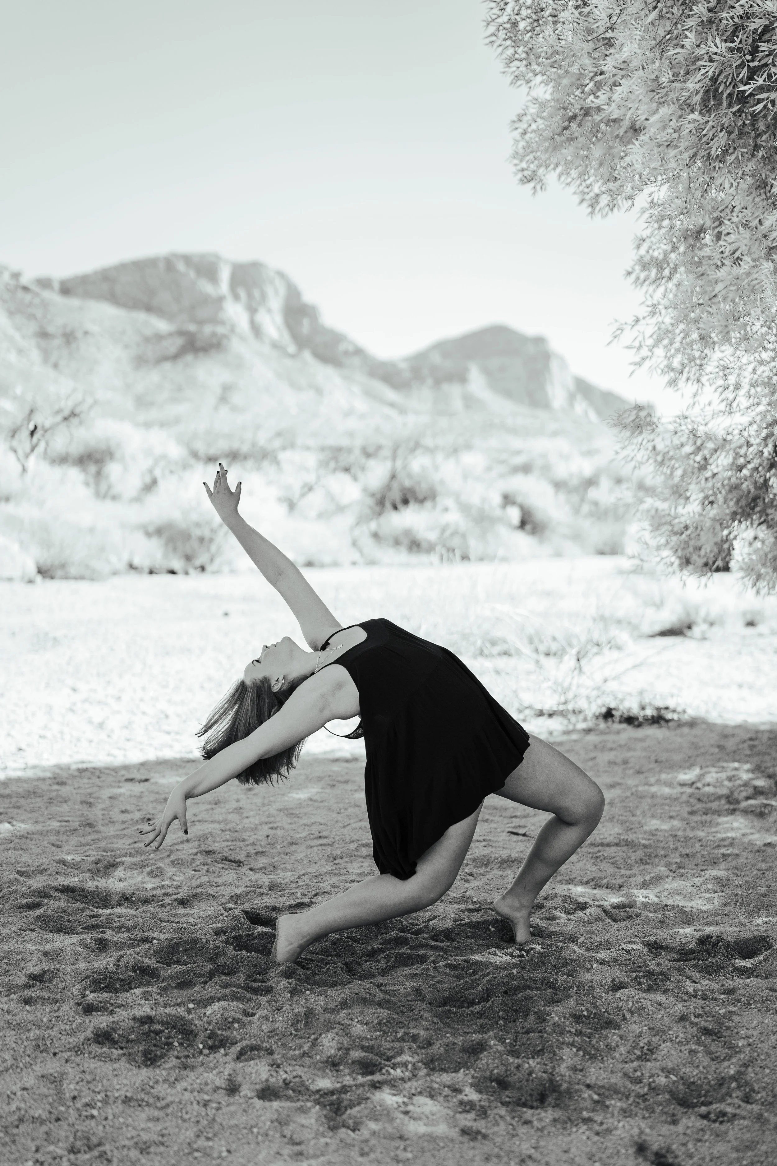 Kayla in a black dress dancing on sandy ground outdoors, with mountains, trees, and a clear sky in the background.