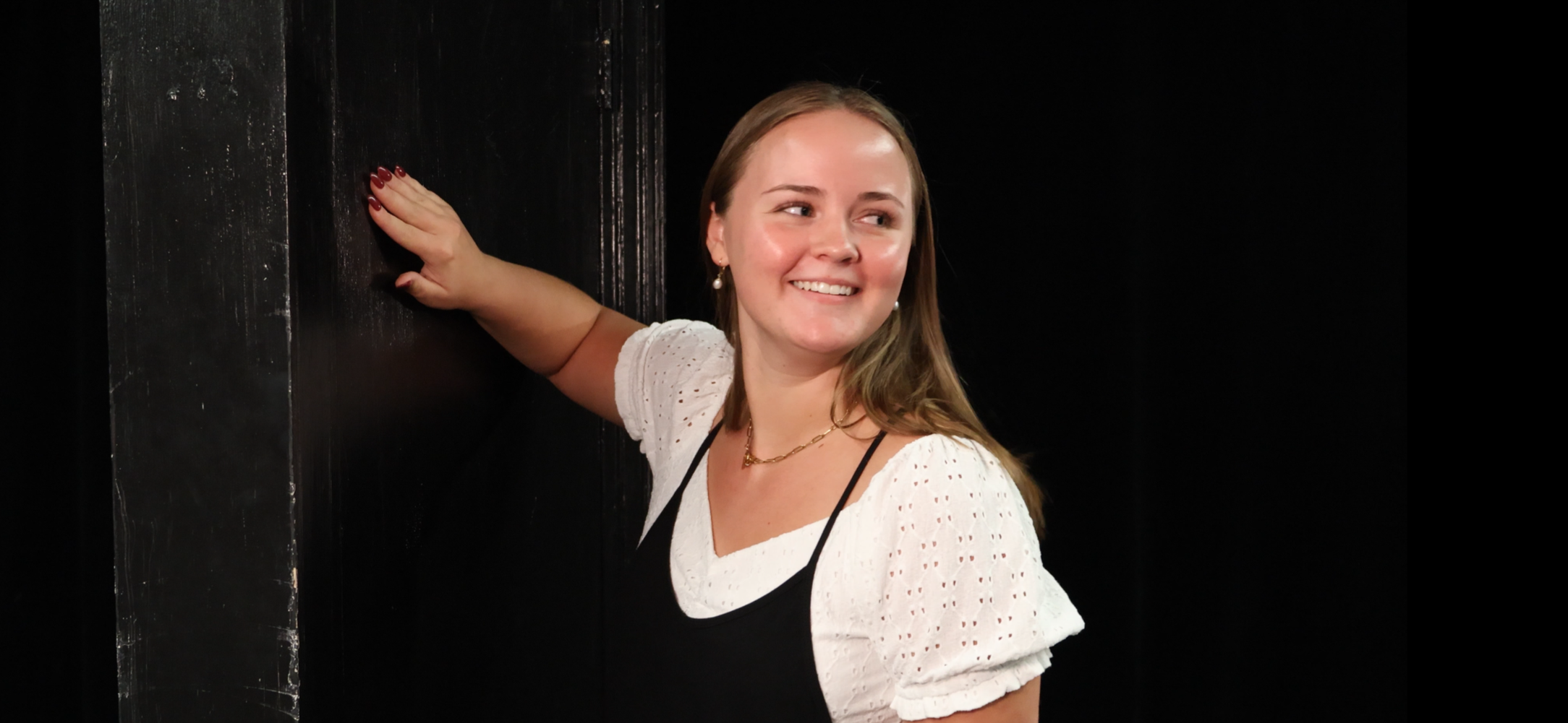 Kayla, woman with long brown hair and earrings, wearing a white blouse and black dress, standing by a black door. Still shot from film project.