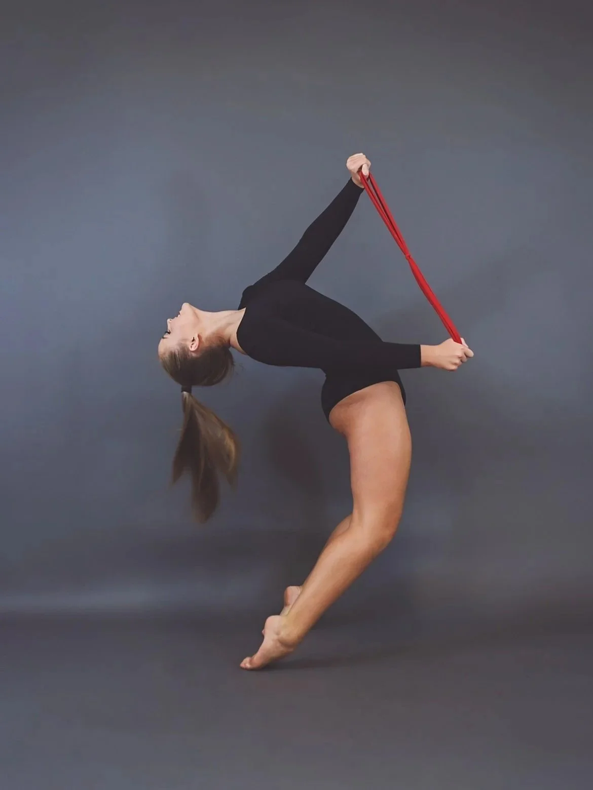Dance photo of Kayla with a red resistance band, bending backward with her arms extended, and her hair in a ponytail, against a plain gray background.