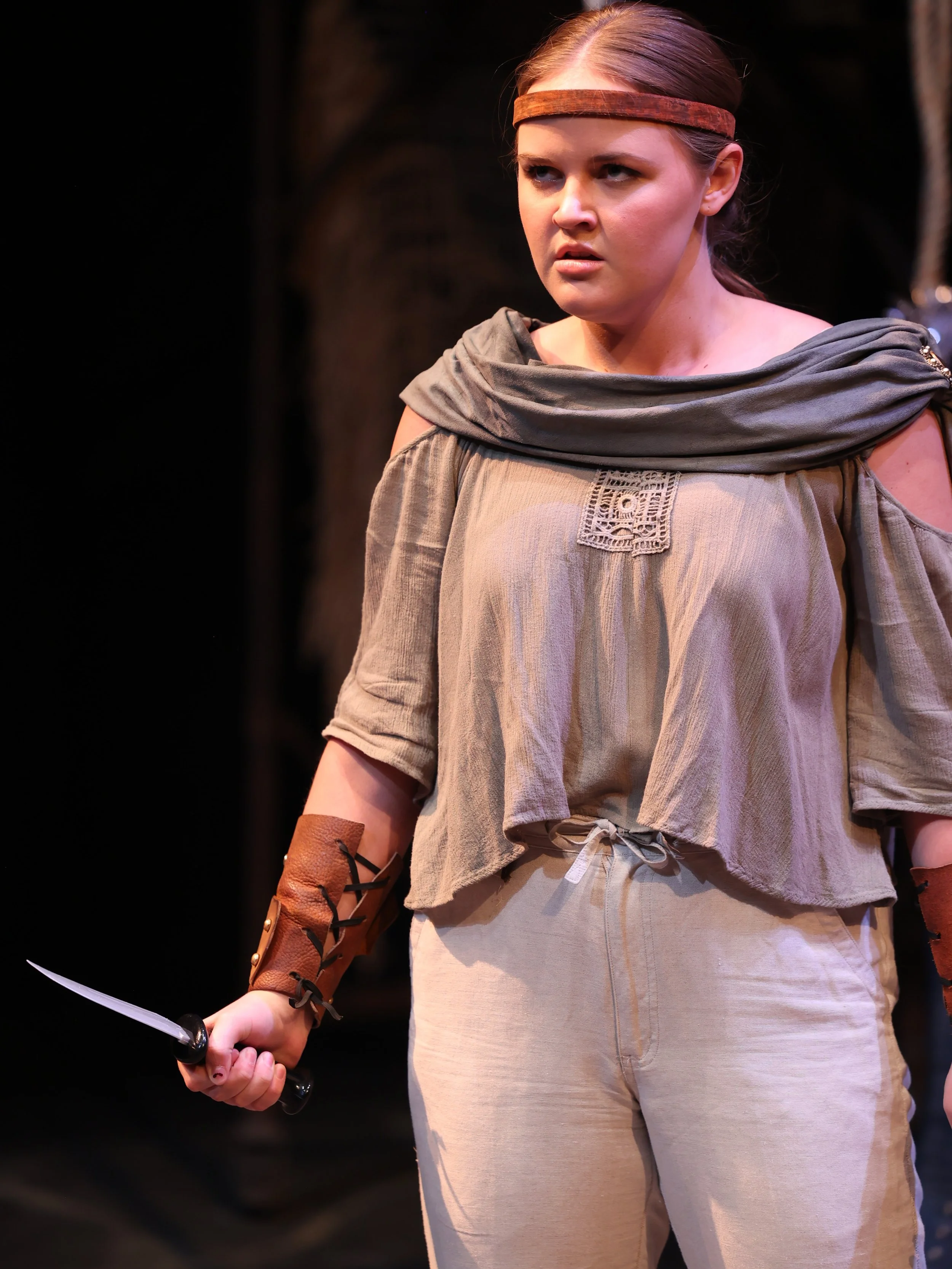 Kayla in a play called Argonautika by Mary Zimmerman, with a focused, angry expression, wearing costumes with earthy tones, a headband, and leather arm guards, holding a small dagger in her right hand, on a stage.