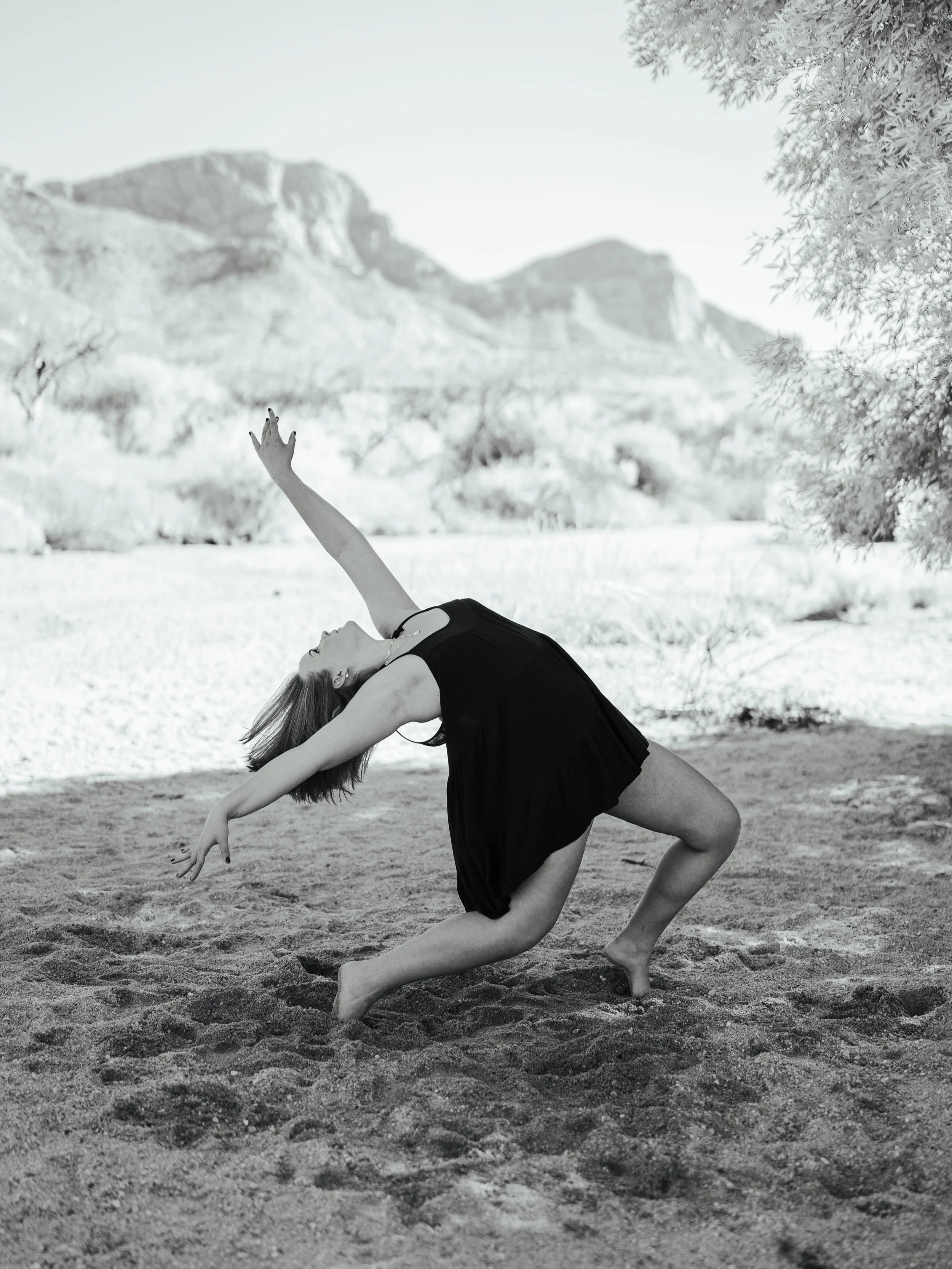 Kayla in a black dress dancing on sandy ground outdoors, with mountains, trees, and a clear sky in the background.
