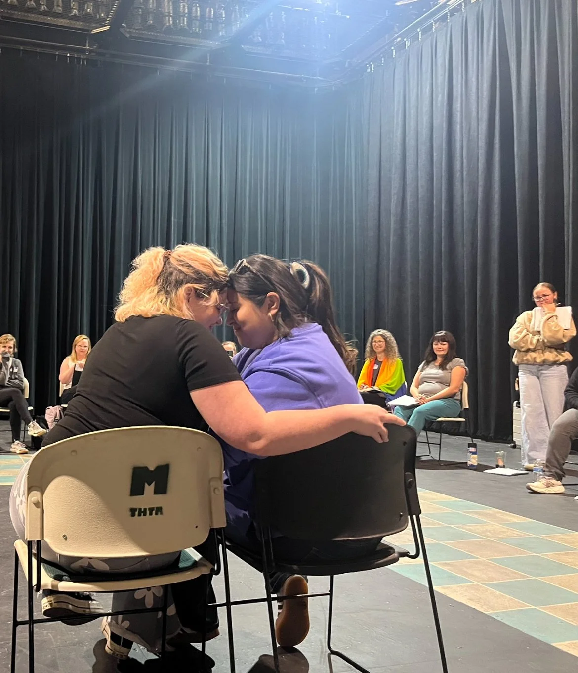 Photo from an Intimacy Choreography workshop, scene choreographed by Kayla Howard. Two performers sit side-by-side in chairs with their foreheads together, with Kayla observing in the back with notebook in tan sweatshirt. 