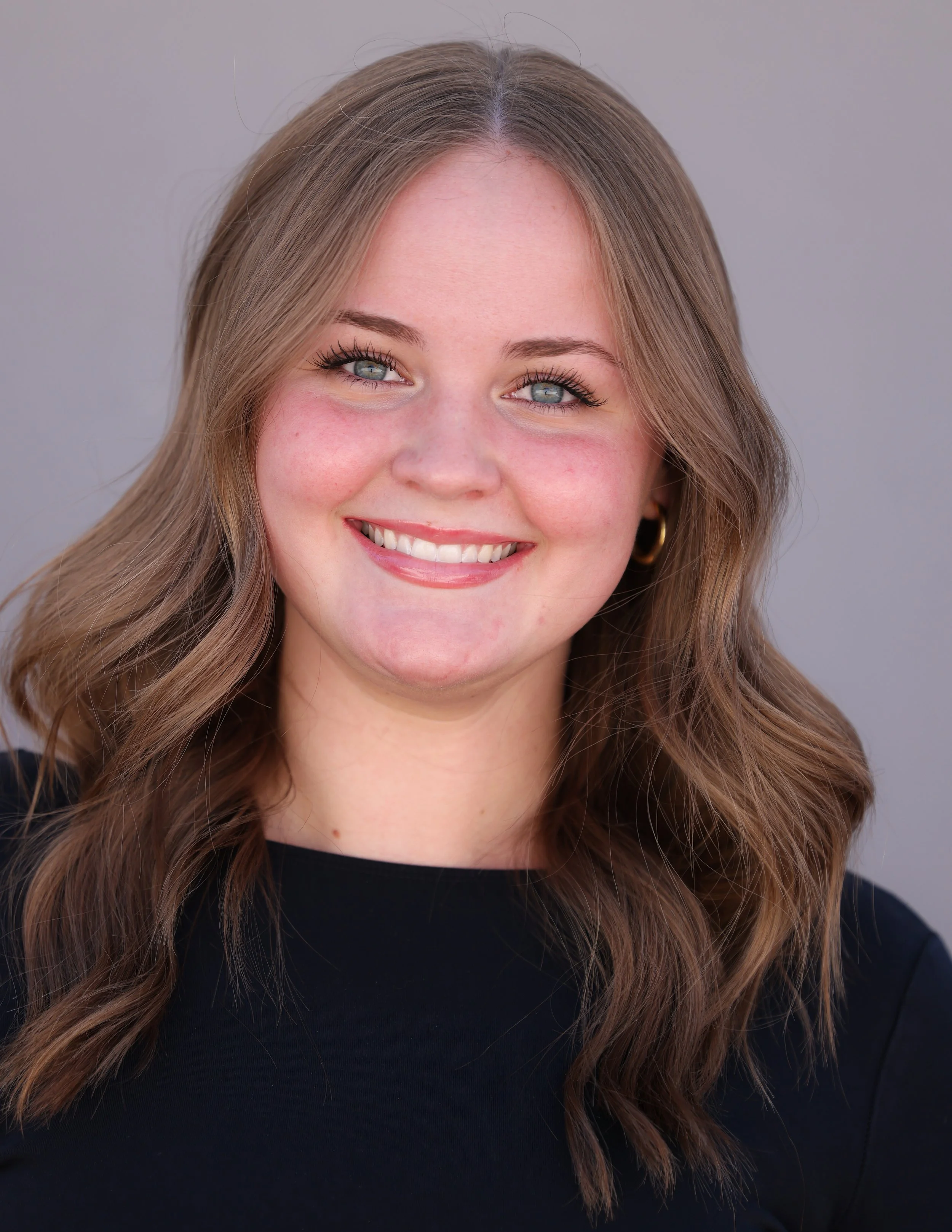 Close-up headshot of Kayla, wavy light brown hair, blue eyes, wearing a black top and gold hoop earrings, smiling against a plain gray background.