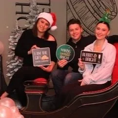 Three people smiling, sitting on a bench in a festive setting, holding holiday-themed signs, with a woman on the left wearing a Santa hat.