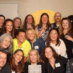 Group of smiling women and one man standing together at a social event, some holding drinks, in a room with a yellow backdrop.