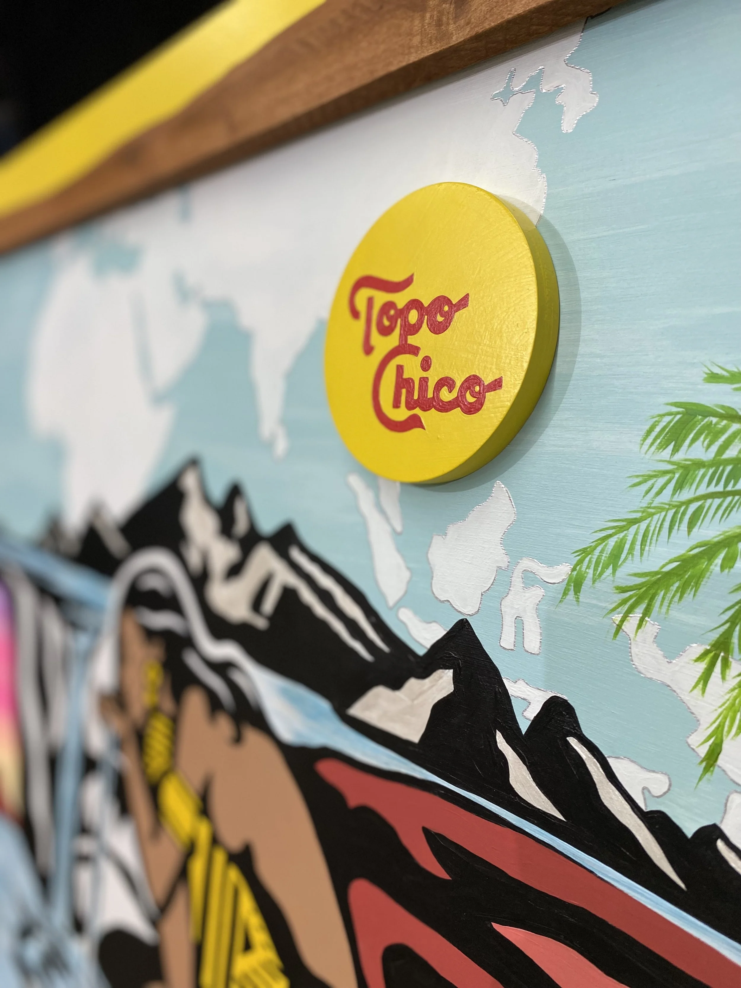 A colorful artwork featuring a map background with the Topo Chico logo in a yellow circle. The map includes mountains and green foliage, with the logo prominently displayed in the upper right portion of the image.