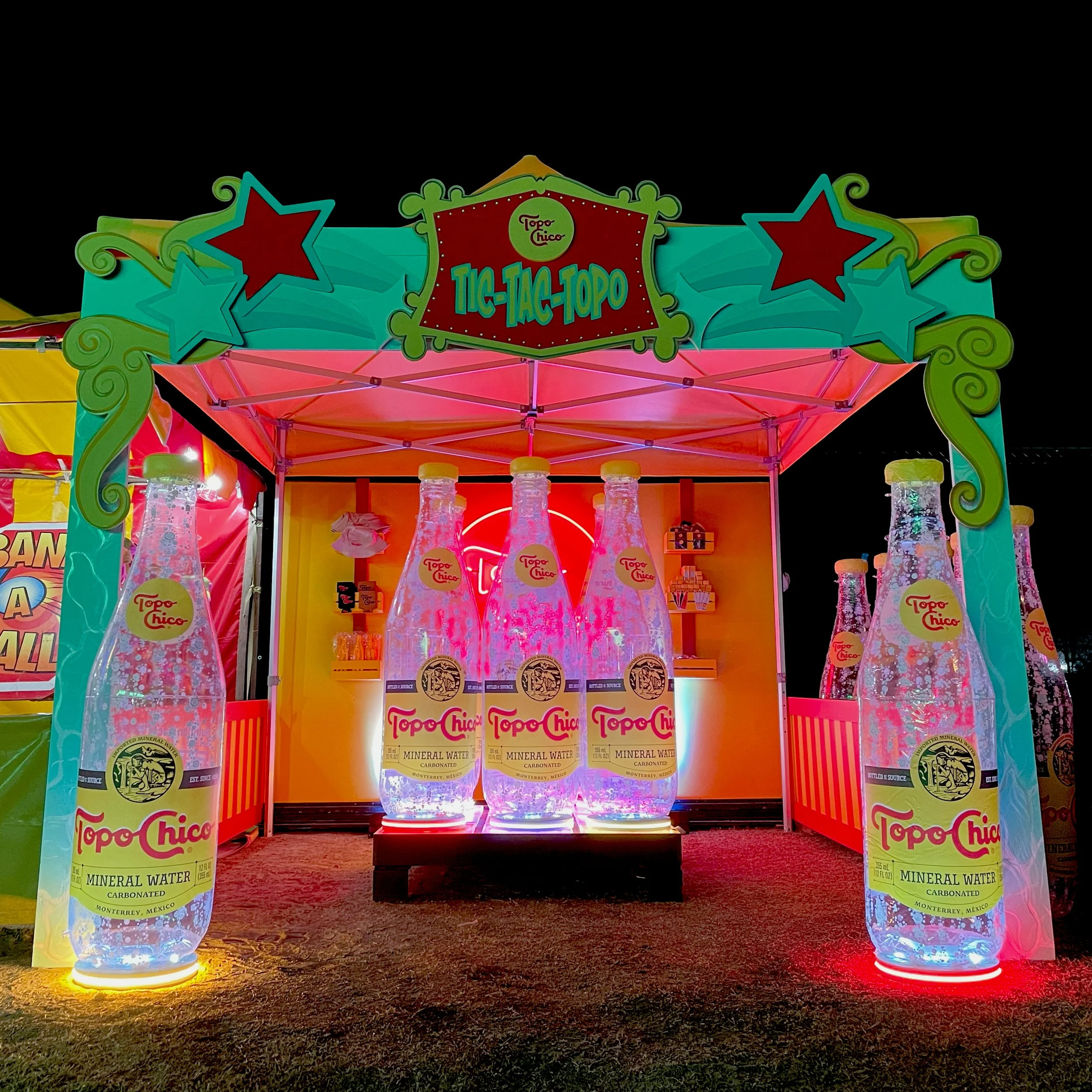 Nighttime carnival booth featuring interactive game with giant, inflatable, bottles of Topo Chico mineral water with glowing lights and colorful signage.