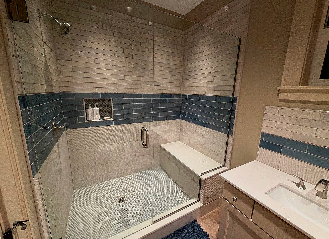 A modern bathroom with a glass-enclosed shower featuring gray and blue tiles, a built-in bench, and wall niche with toiletries, alongside a white vanity with a sink and faucet.