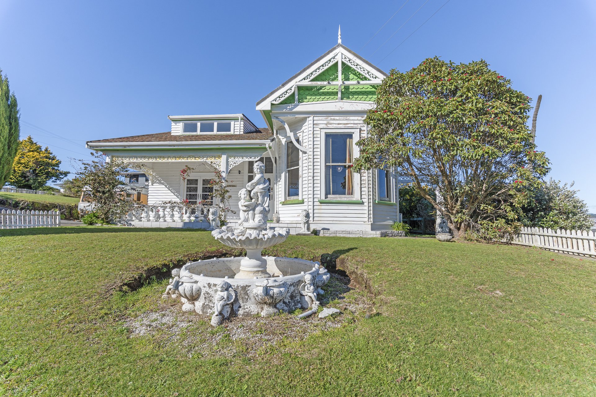 265f Tukapa Street, Westown, New Plymouth. A grand Victorian-style white house with green accents, surrounded by a well-kept lawn, trees, a white picket fence, and a decorative white fountain with cherub statues in the front yard.