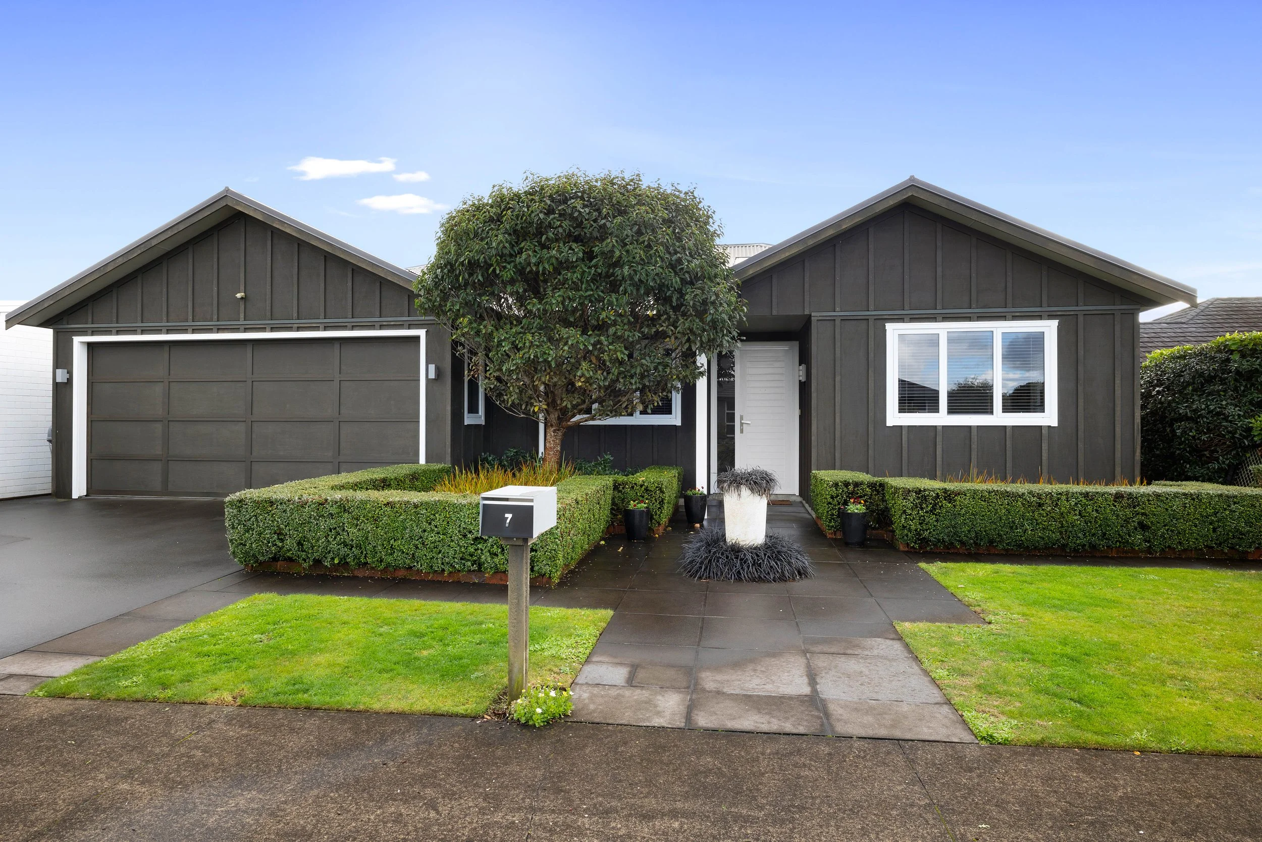7 Gleneagles Way, The Links, Waiwhakaiho, New Plymouth.  Front view of a modern, dark brown board and batten house with white trim, a driveway, and a well-maintained lawn and garden.