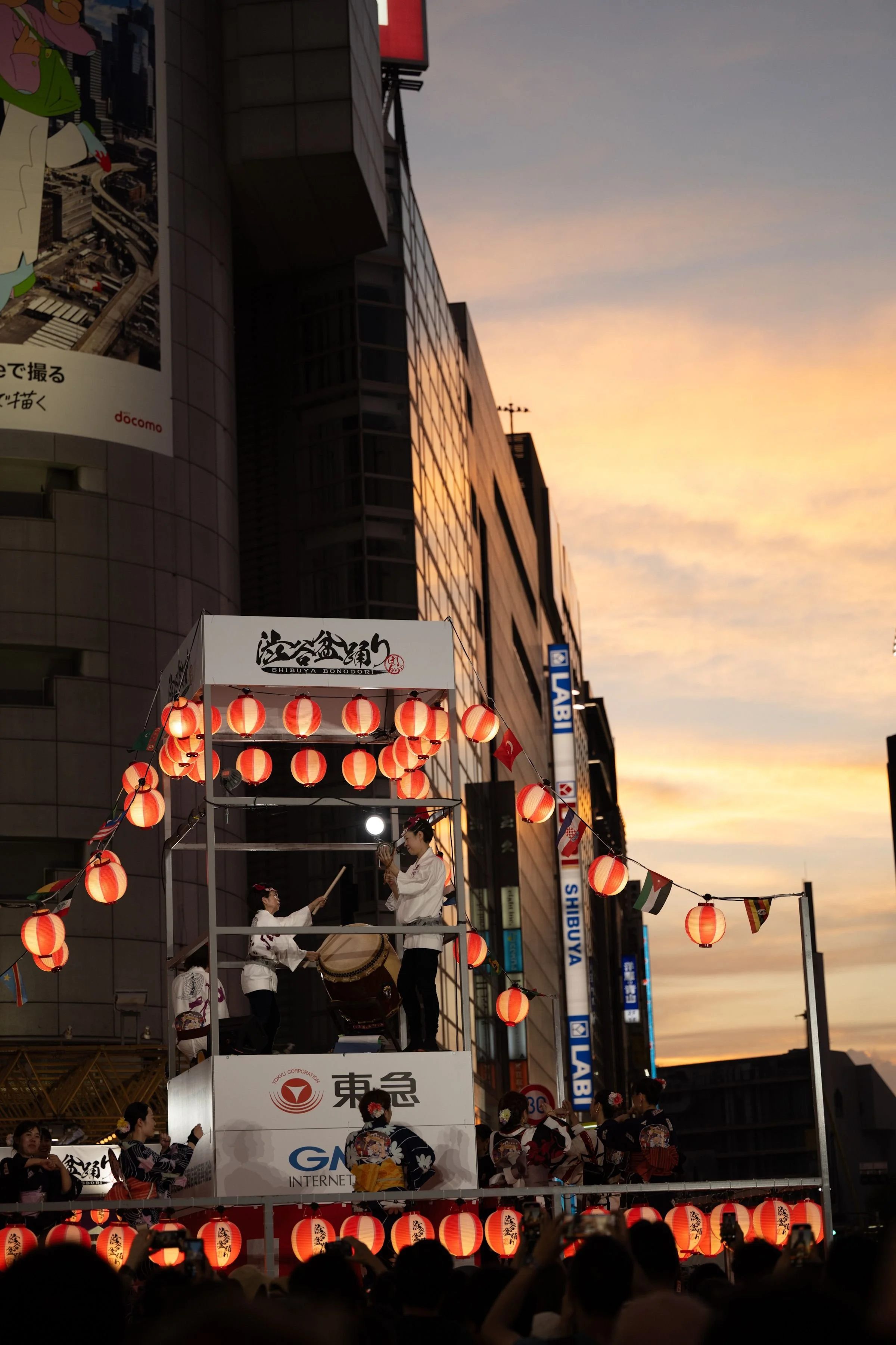 A stage is at the center of circle. On the stage people are playing traditional Japanese drums and around the stage people wearing Yukatas are dancing to show the crowd the choreography to follow. The stage is surrounded by red and white lanterns.