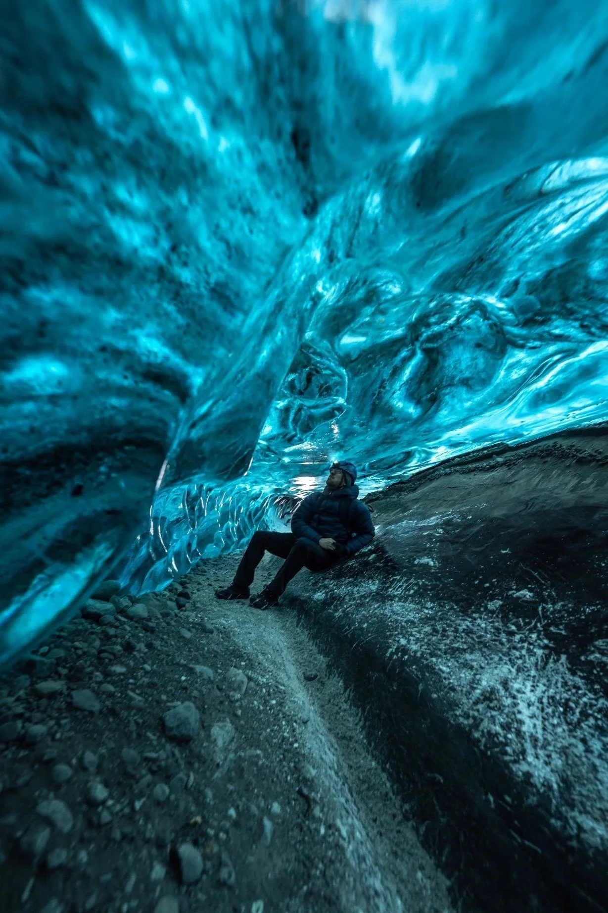 A person sitting inside an ice cave with blue ice formations overhead and rocky ground beneath.