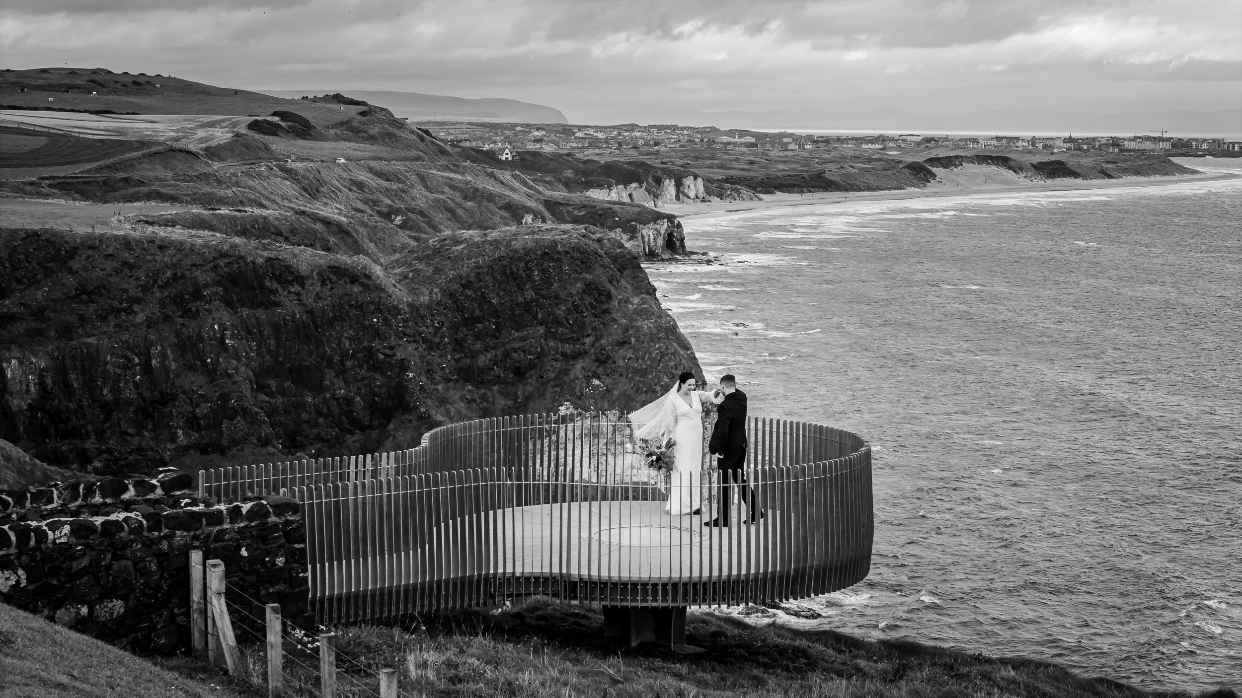A black and white photo of a bride and groom standing on a circular platform with a railing, overlooking the ocean near cliffs, during a wedding ceremony.