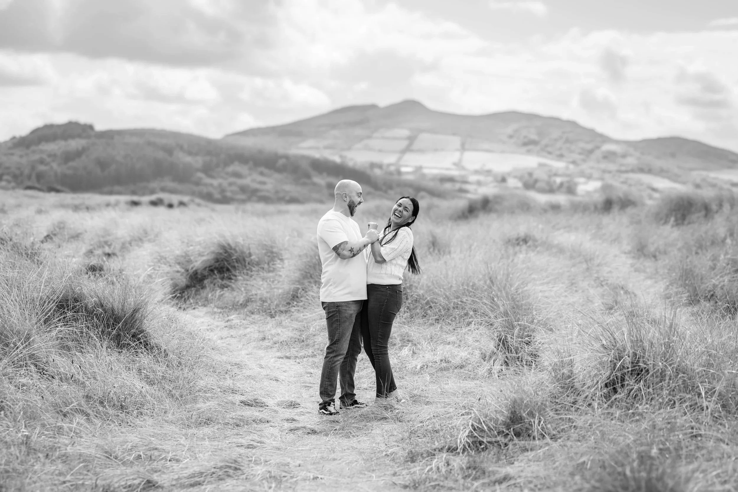 A black and white photo of a happy couple standing on a dirt path in a grassy field, laughing and holding each other's hands with mountains in the background under a partly cloudy sky.