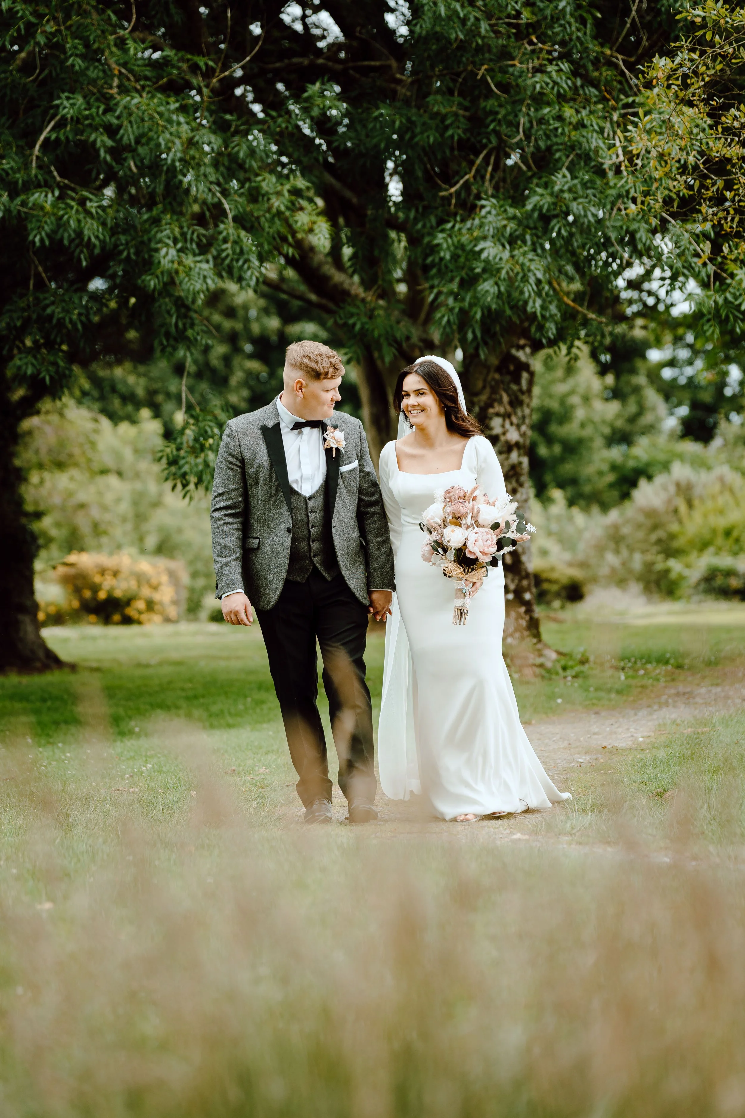 A bride and groom walking hand in hand outdoors, dressed in wedding attire, with trees and greenery in the background.