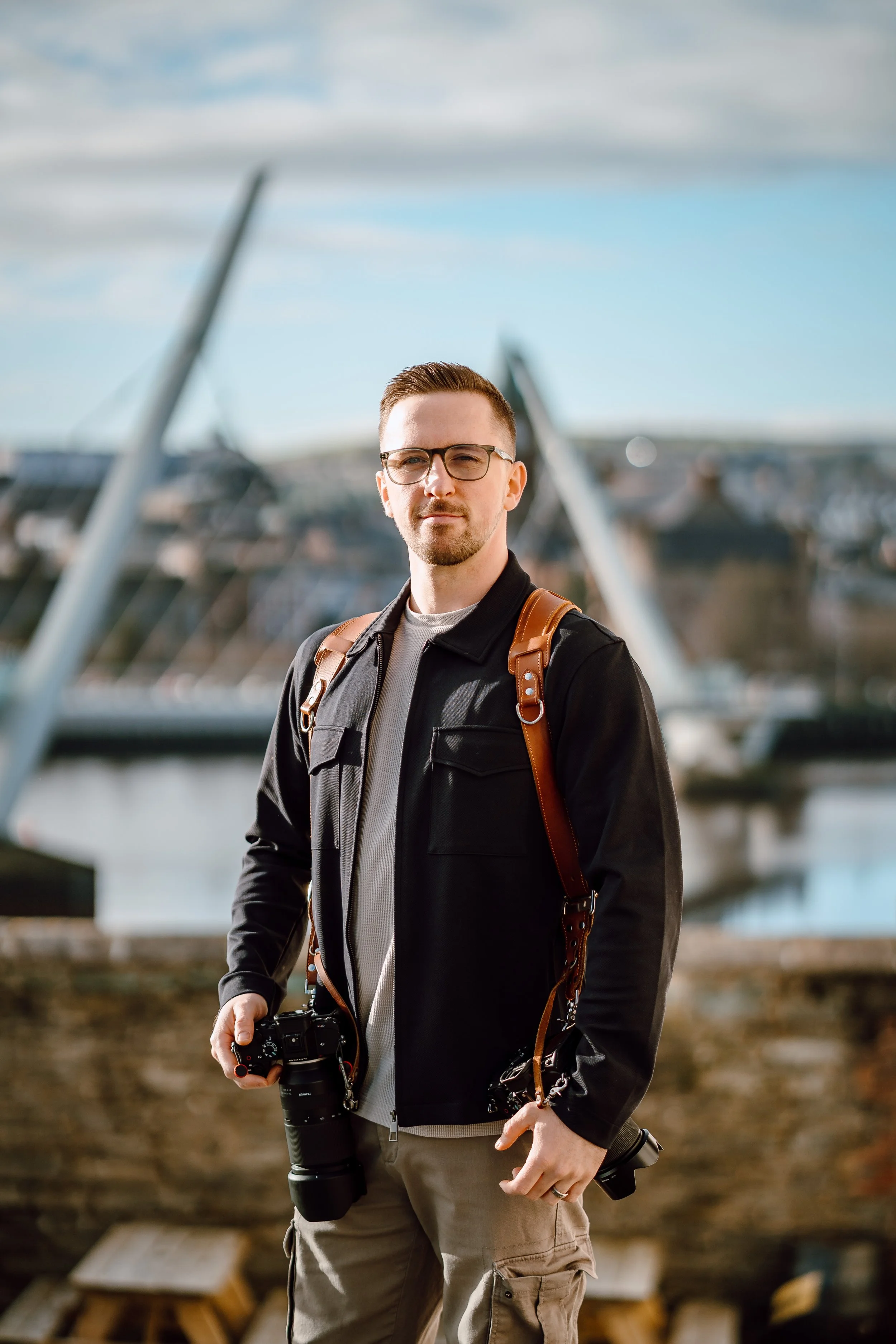 A man with glasses holding a camera standing outdoors near a bridge, wearing a black jacket and carrying a backpack.