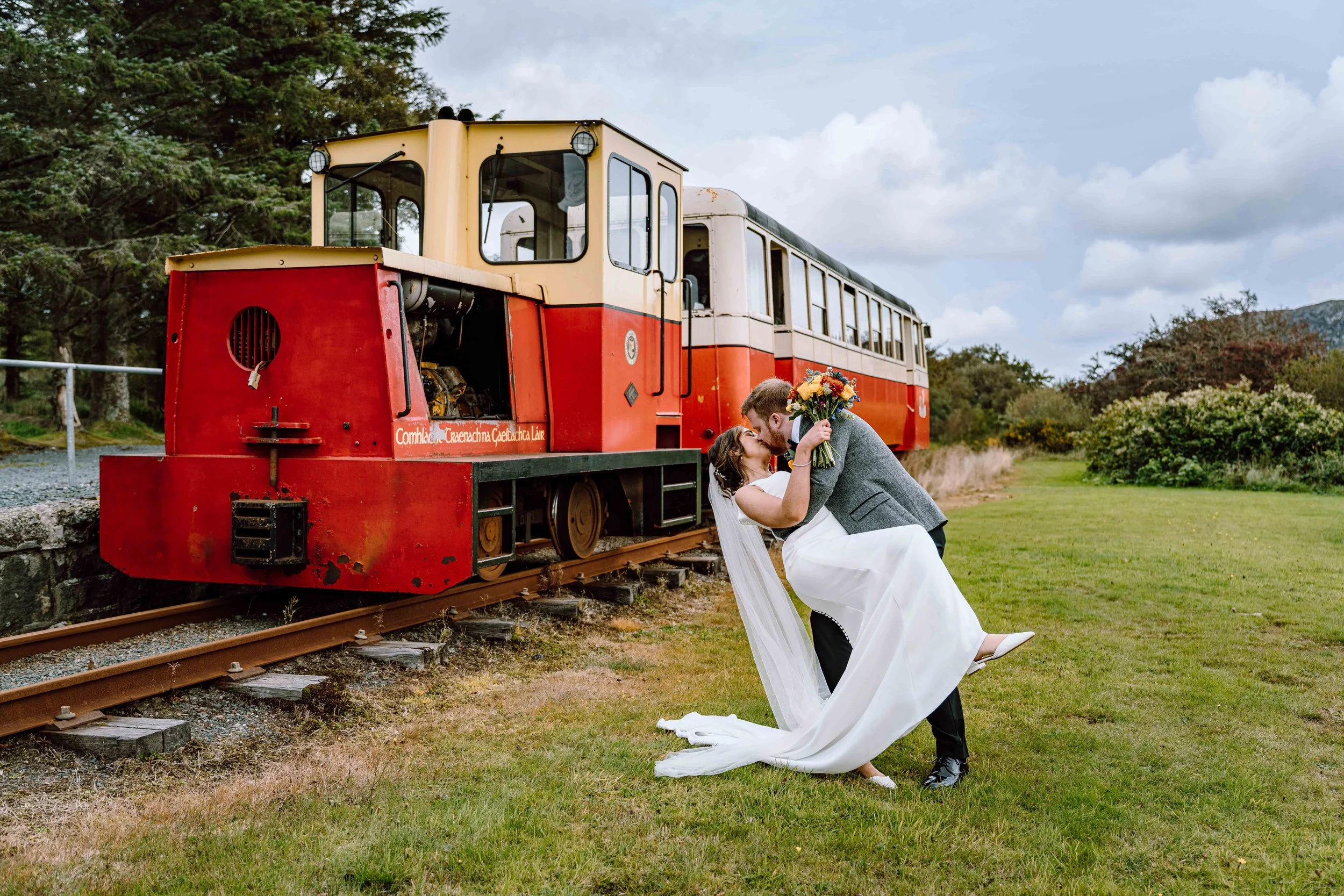 A newly married couple in wedding attire sharing a kiss outdoors near a small vintage train engine on the tracks. The groom dips the bride, holding a bouquet of flowers, against a backdrop of trees and a cloudy sky.