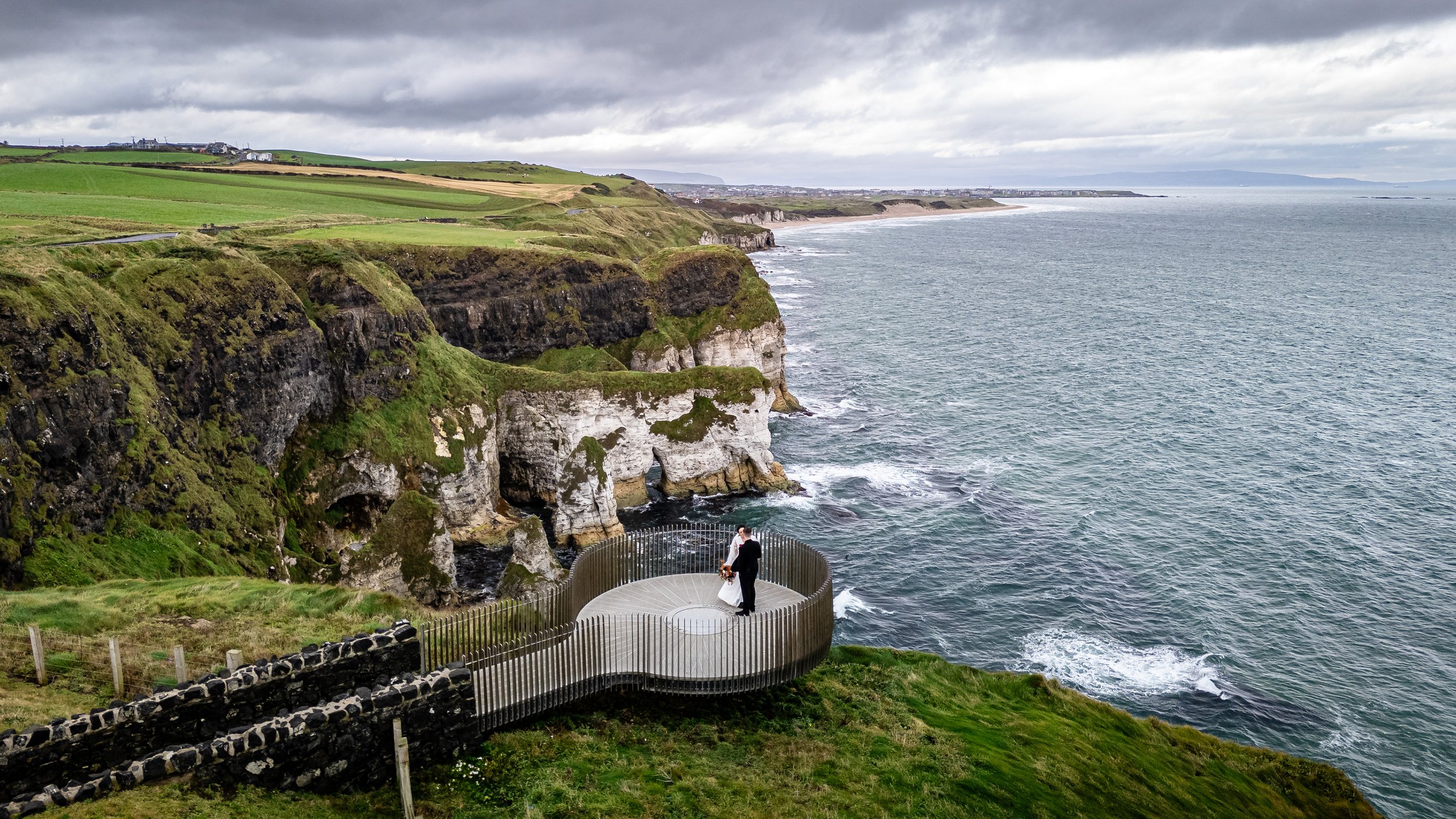 A couple stands on a circular viewing platform on a cliff overlooking the ocean, with green fields and rocky cliffs in the background under a cloudy sky.