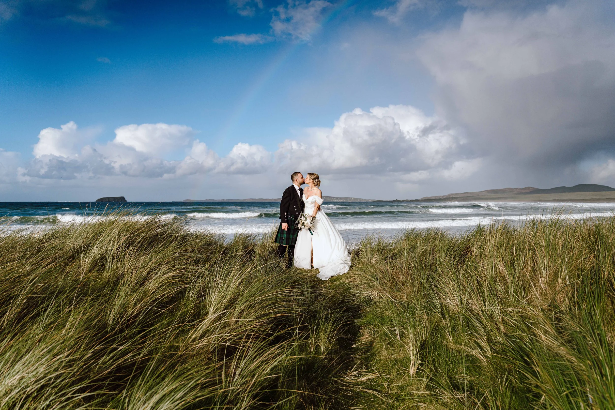 A bride and groom standing on a grassy dunes overlooking the ocean, sharing a kiss on their wedding day with a partly cloudy sky and distant islands in the background.