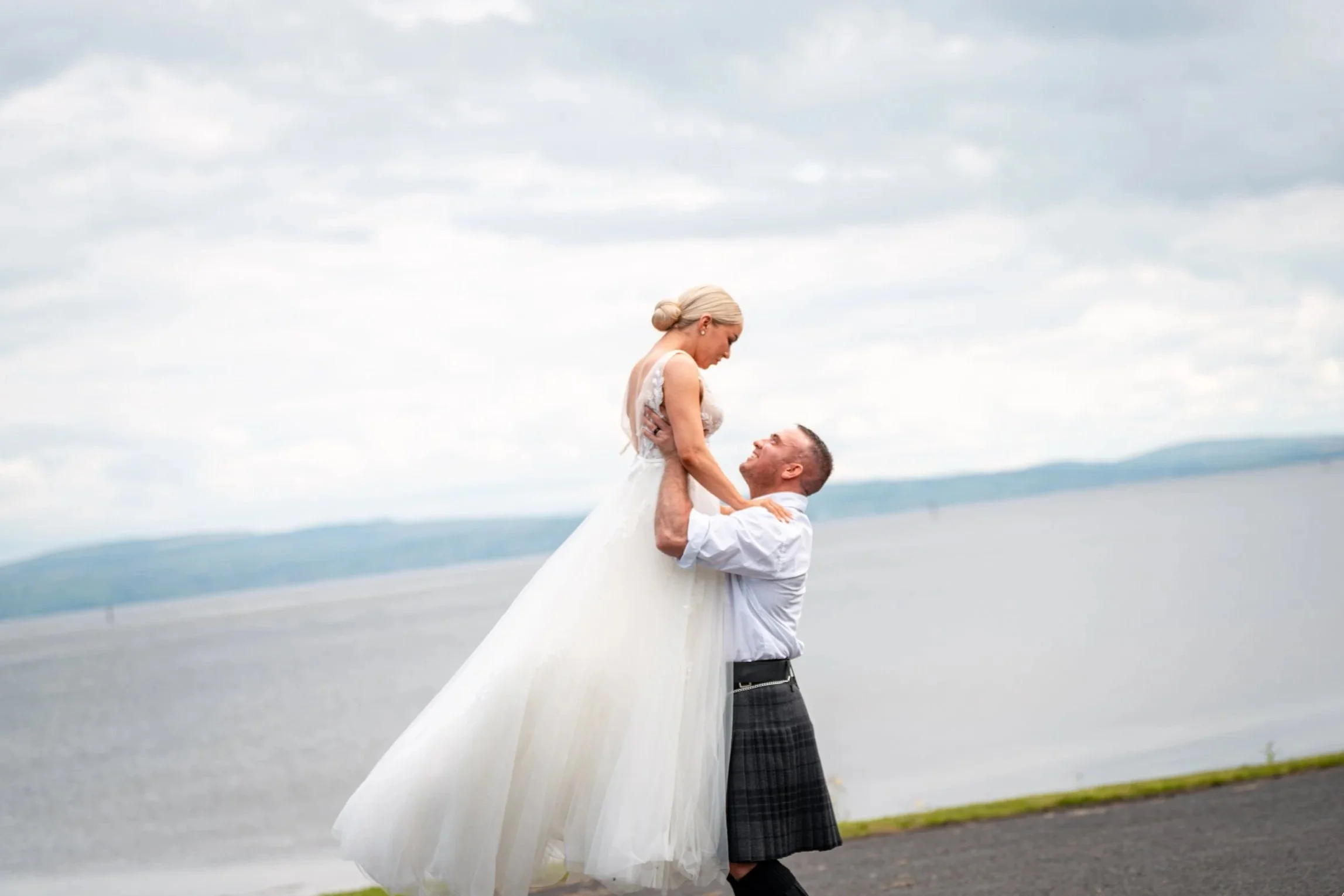 A man in traditional Scottish kilt lifts a woman in a white wedding dress against a lakeside background with clouds.