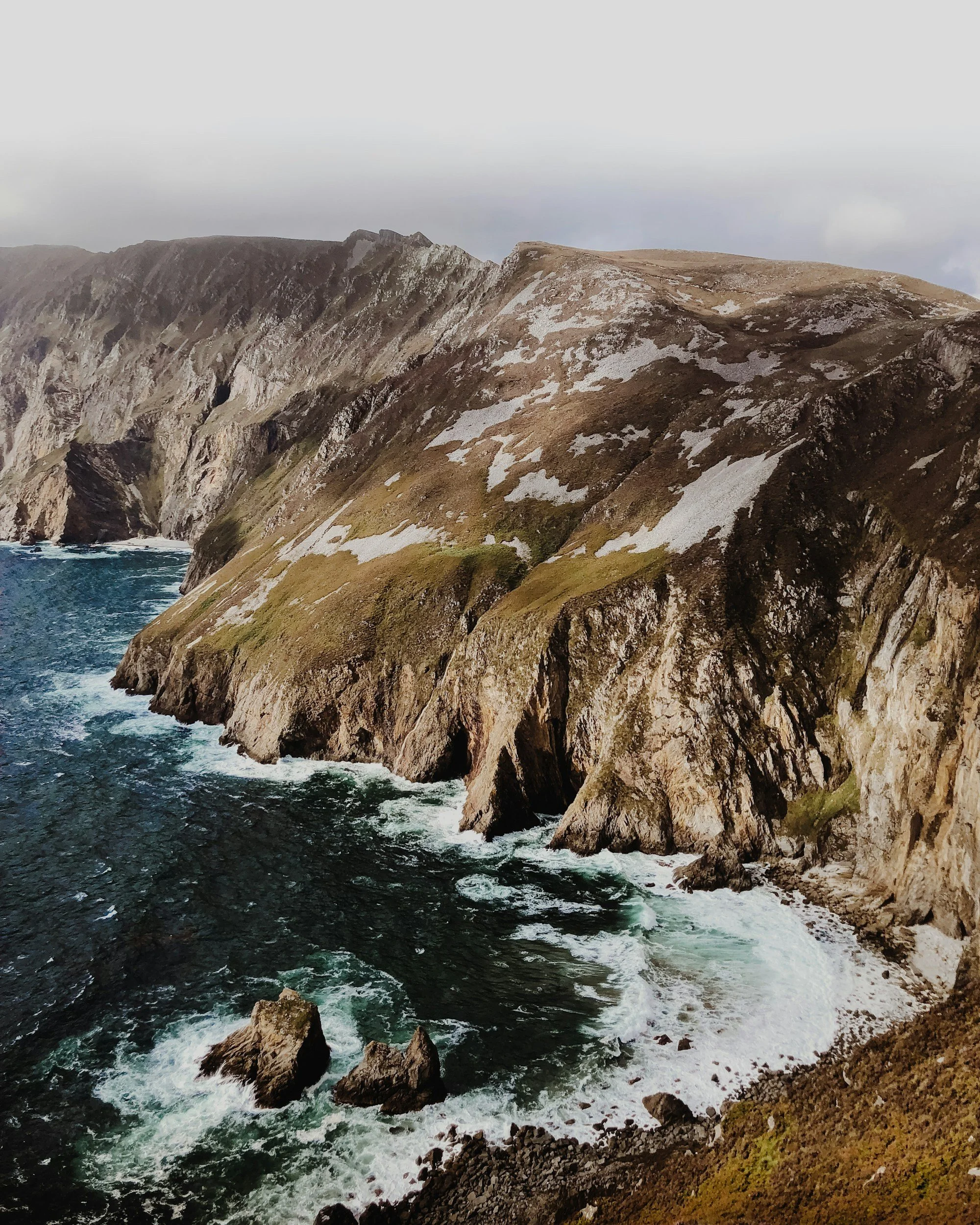 Cliffside coastline with rocky cliffs and ocean waves crashing at the base, some patches of snow on the cliffs.