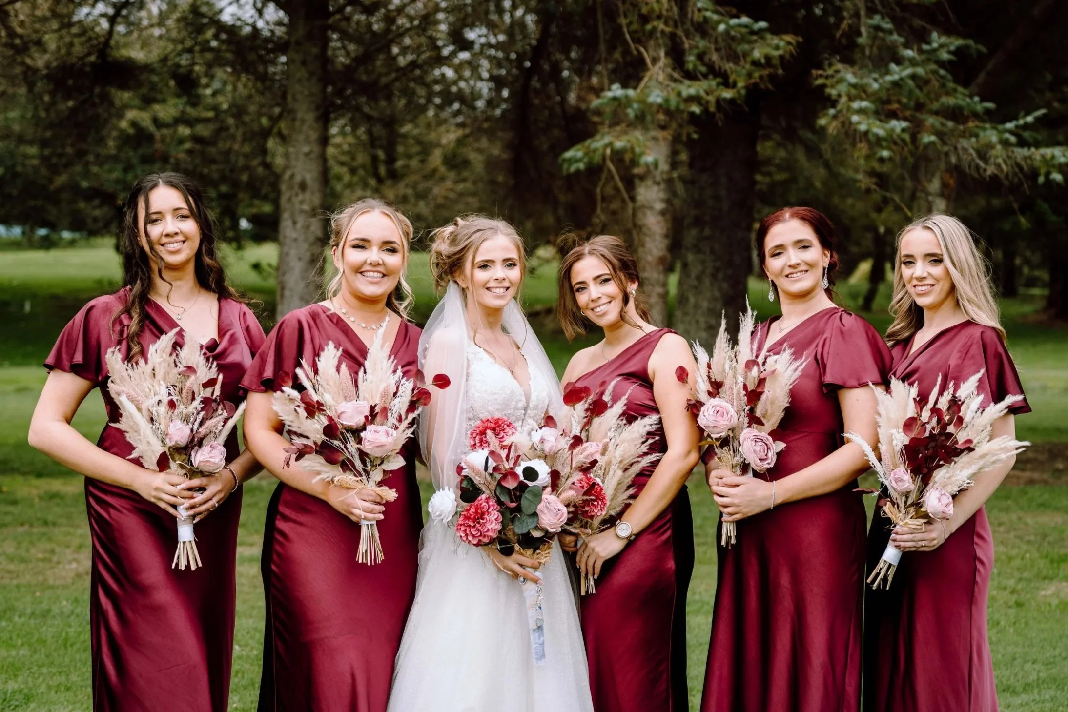 A bride with five bridesmaids standing outdoors in a park with trees in the background. They are all smiling and holding bouquets of pink and white flowers with pampas grass. The bridesmaids are wearing matching maroon dresses, and the bride is weari