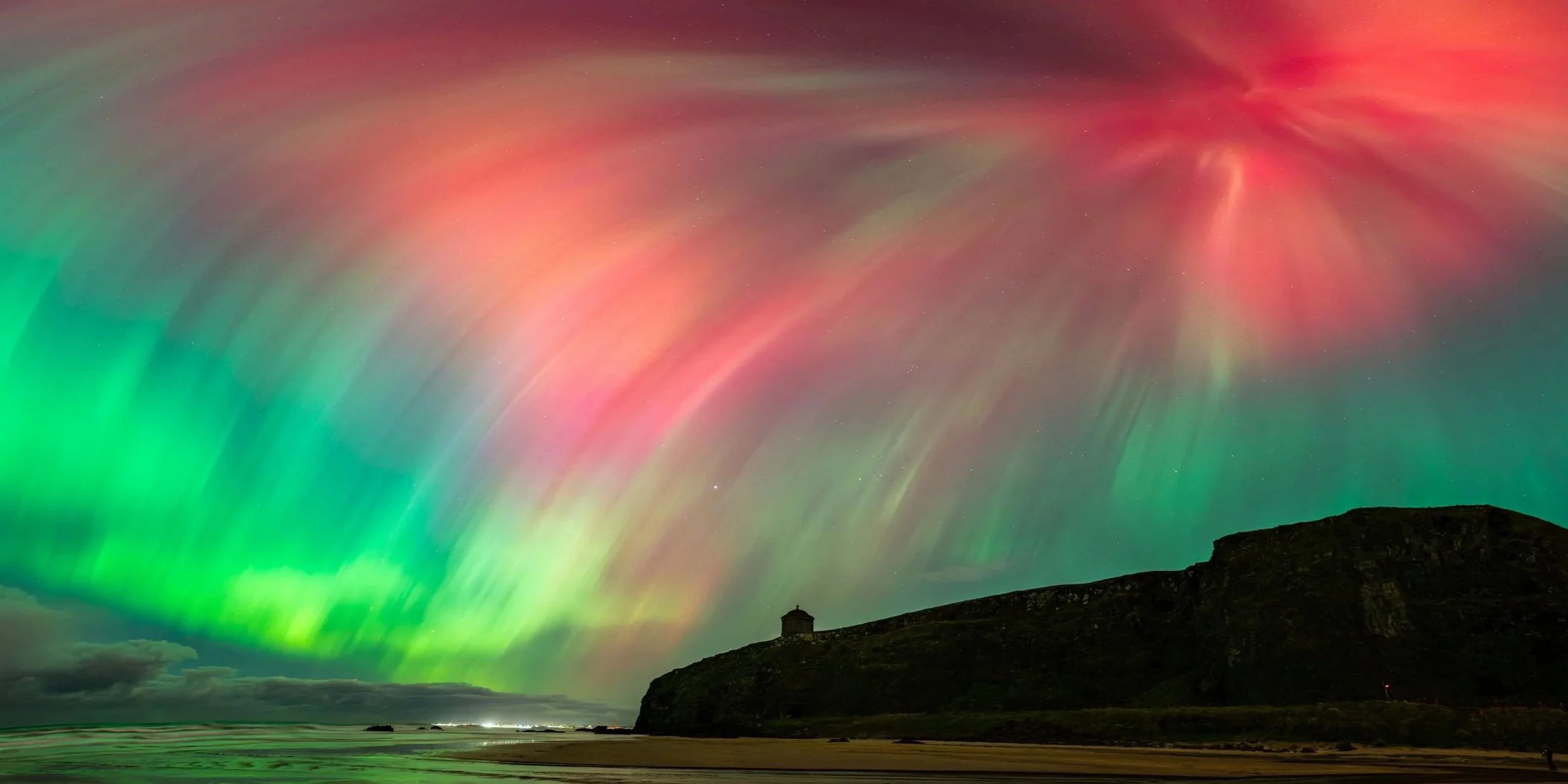 Northern lights over a beach with a small pavilion on a hill, dark clouds near the horizon, and rocky formations in the distance.