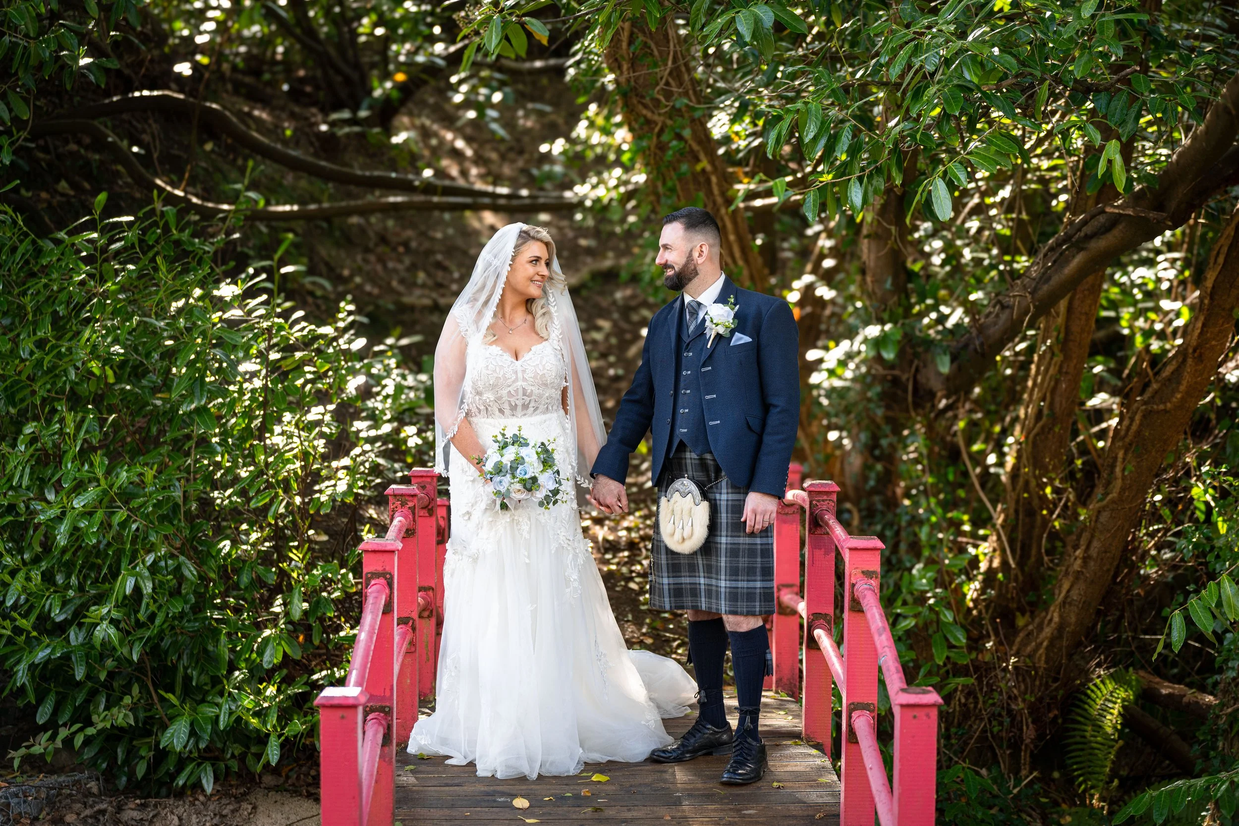 A bride and groom standing on a small red bridge in a lush green forest, holding hands and looking at each other during their wedding photos.