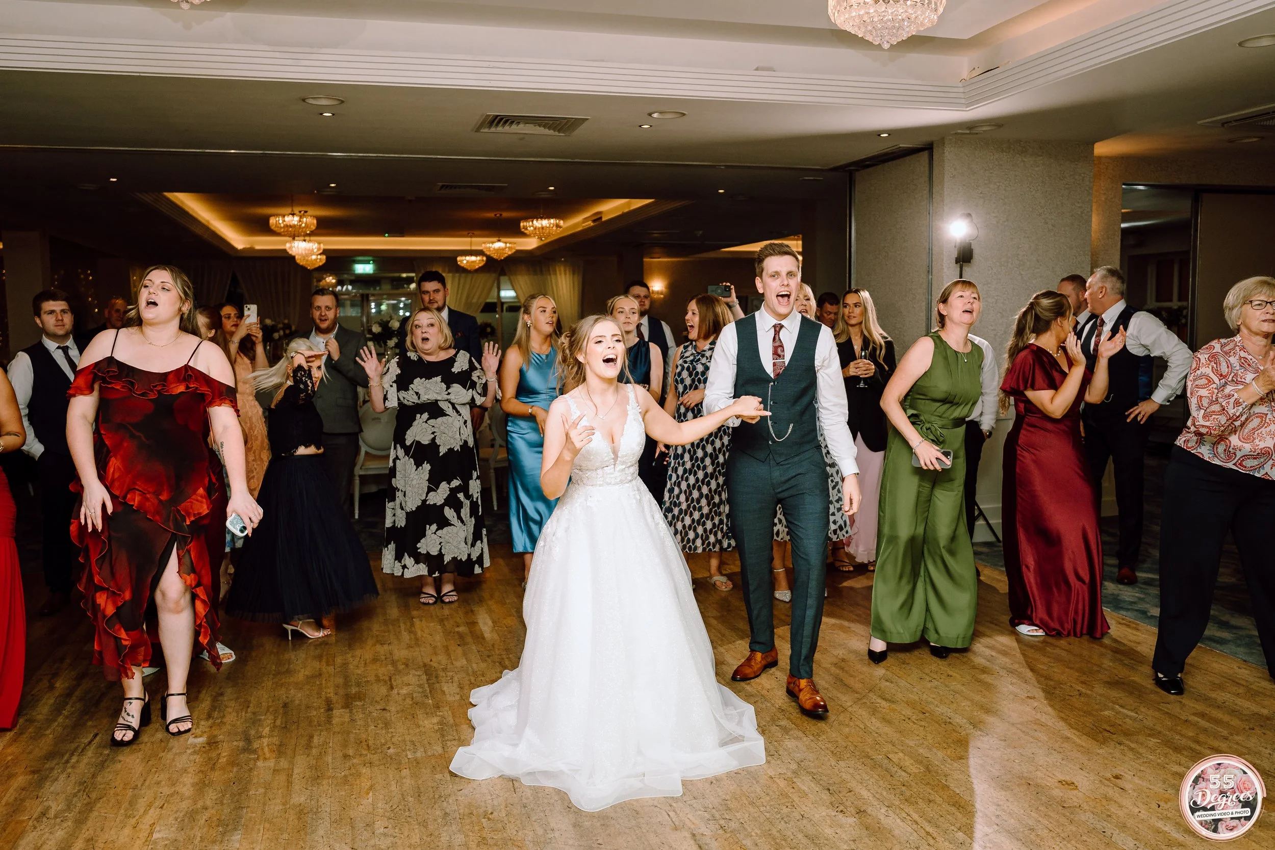 People dancing and celebrating at a wedding reception, with a bride in a white wedding dress and a groom in a suit at the center.