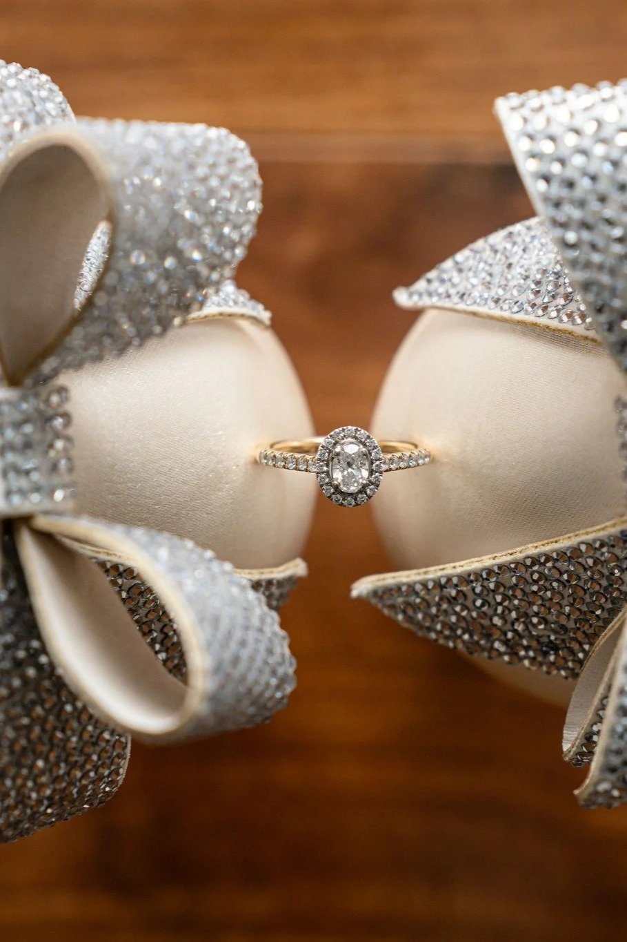 Close-up of a diamond engagement ring with an oval-shaped center stone surrounded by smaller diamonds, placed between two cream-colored cushioned rings and decorated with a sparkly, rhinestone-encrusted bow.