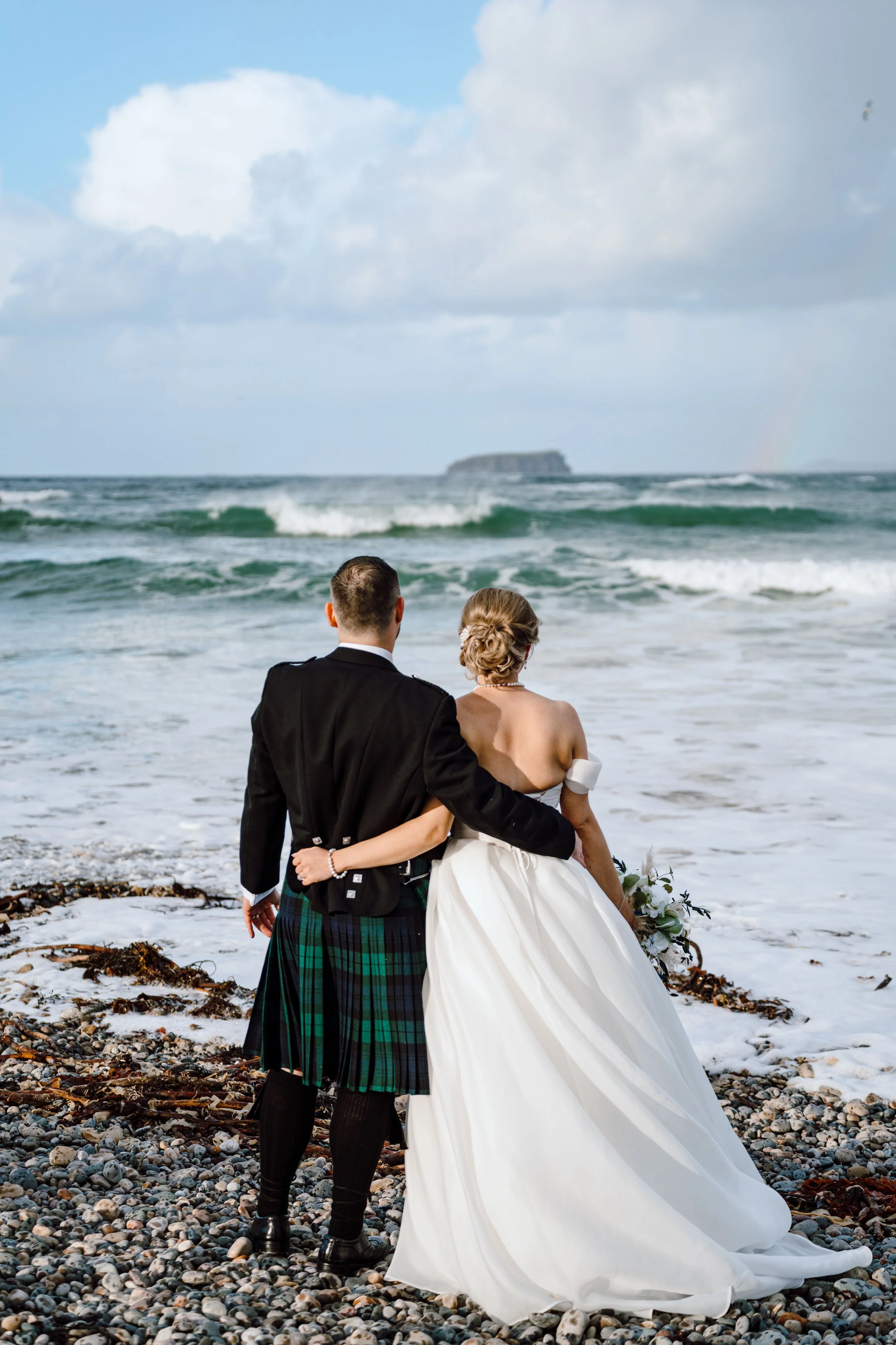 A newlywed couple stands on a rocky beach, facing the ocean, with an island in the distance and a rainbow in the sky.