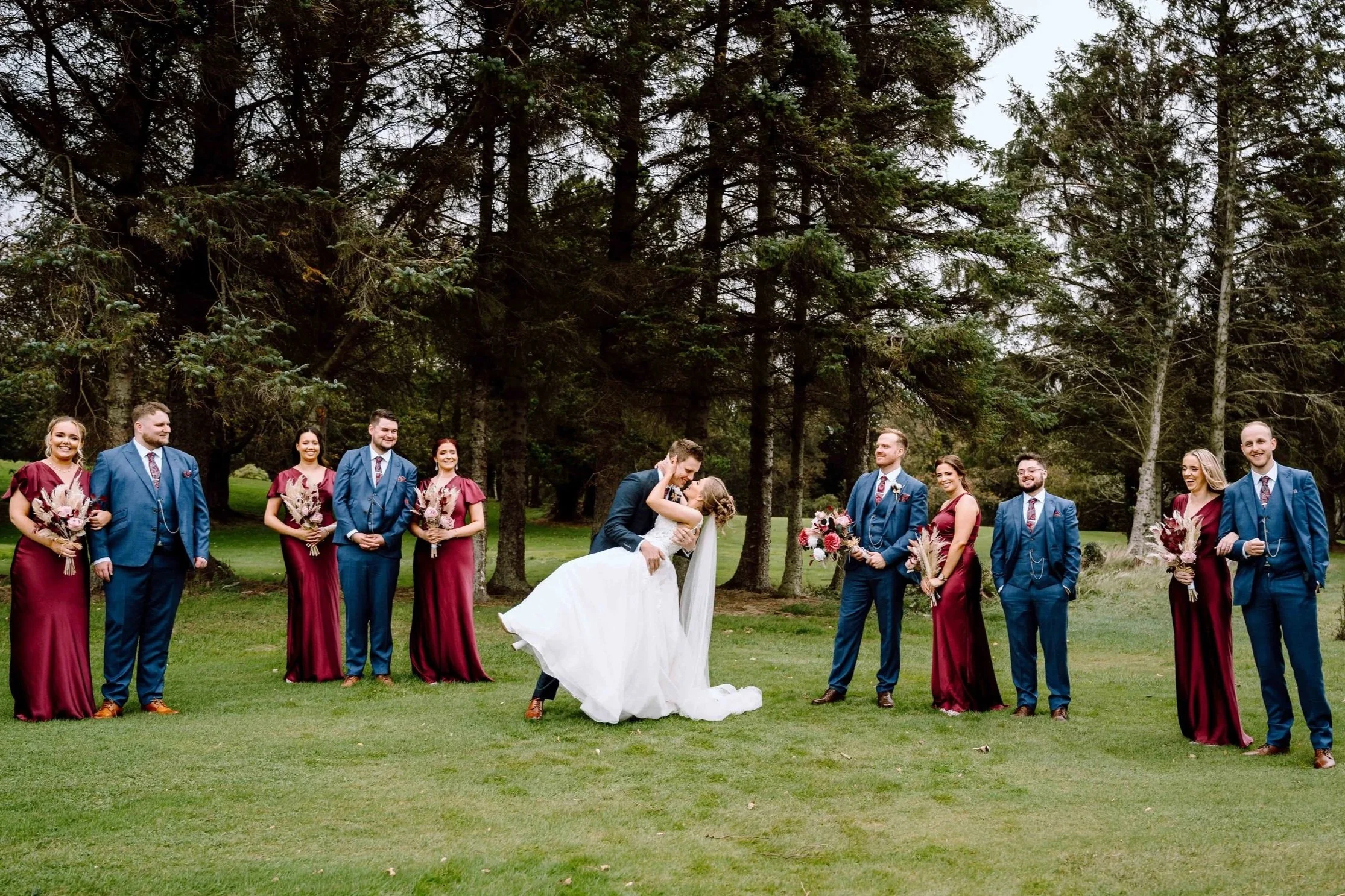 A wedding party outdoors in a wooded area: the groom dips and kisses the bride, who is in a white wedding dress, while her groom kisses and dips her. The bridal party stands on both sides, with women in matching burgundy dresses and holding bouquets, and men in blue suits, some holding flowers, smiling and looking at the couple.