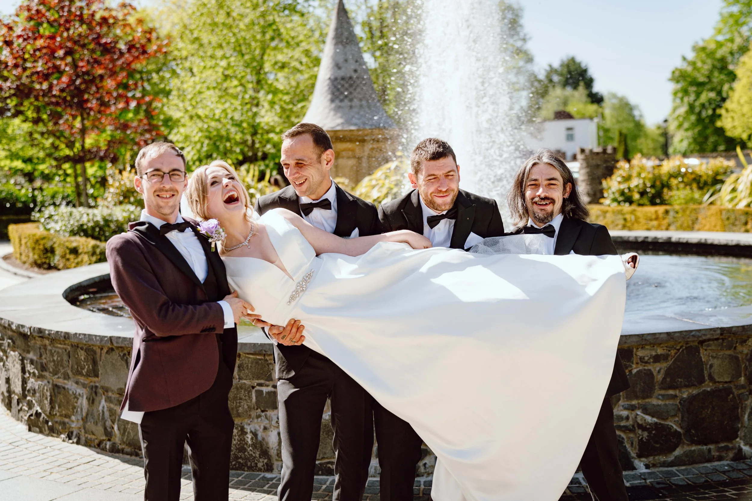Bride in a wedding dress being held up by groom and groomsmen at a park with a fountain in the background.
