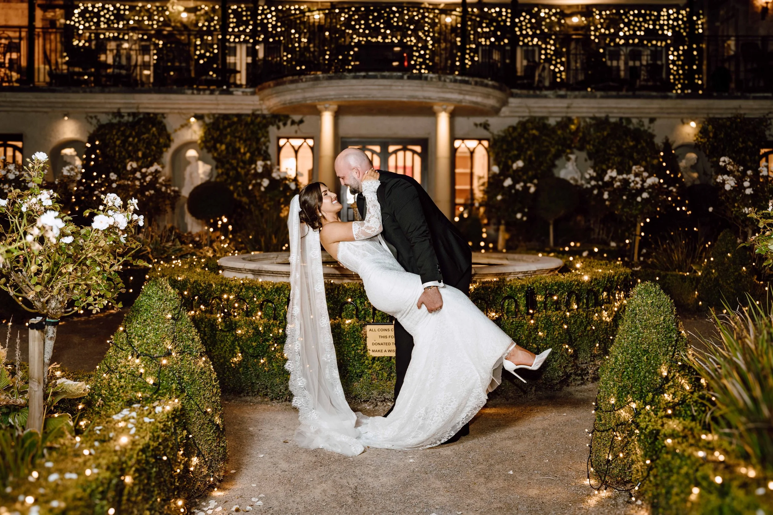 A bride and groom dancing at night in front of a lit fountain at a wedding celebration, surrounded by illuminated greenery and string lights.